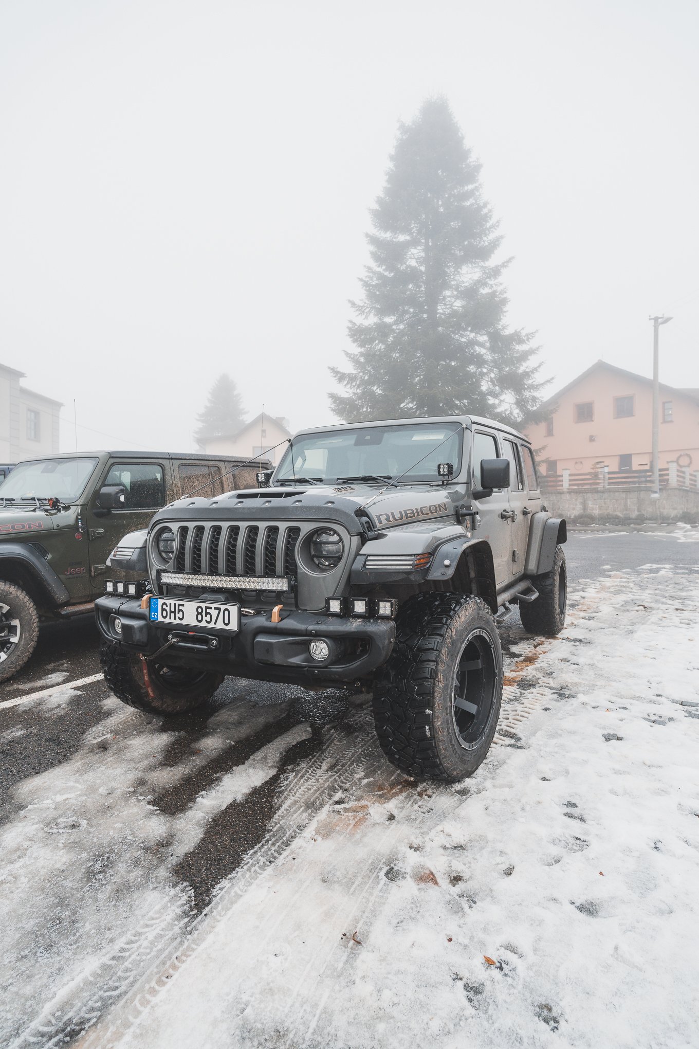 A black off-road Jeep Rubicon parked on a snowy and icy surface in a foggy outdoor area, with another vehicle partly visible next to it, and trees and buildings in the background.