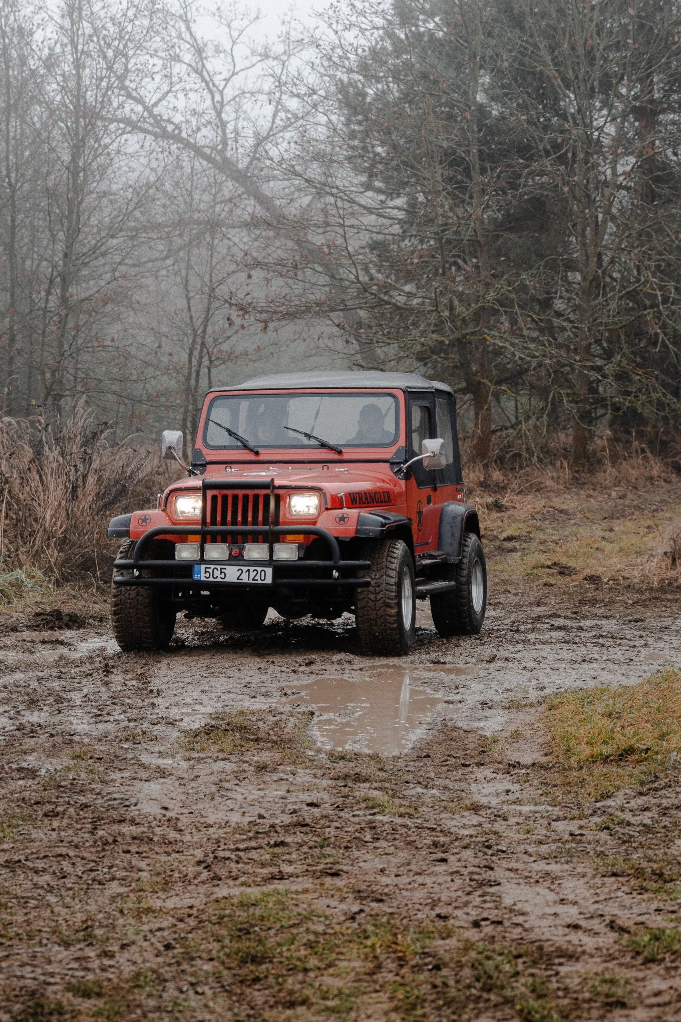 Red Jeep Wrangler on a muddy dirt path in a foggy, leafless forest.