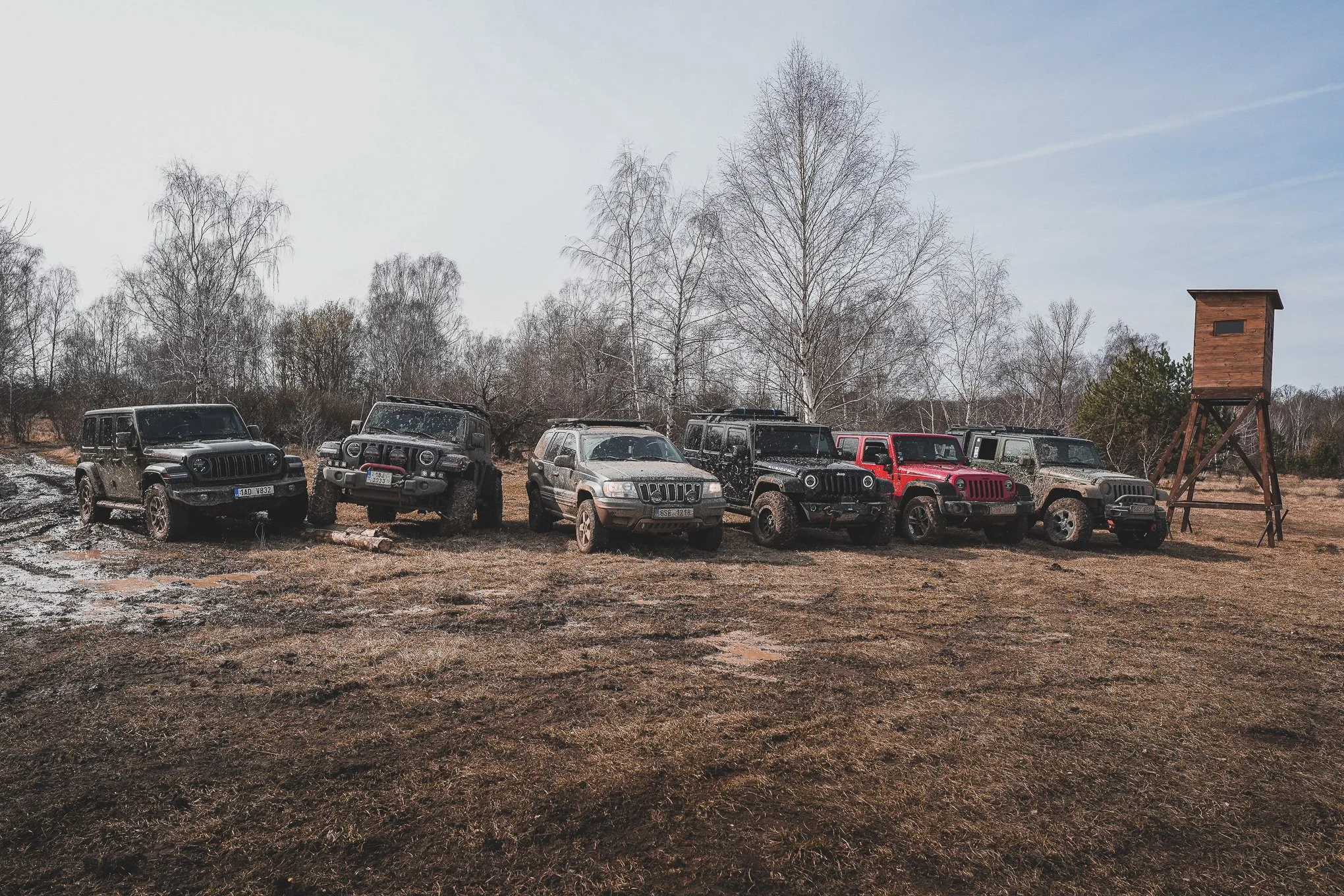 Seven Jeep vehicles parked on a muddy field during daytime, with leafless trees and a wooden hunting blind in the background.