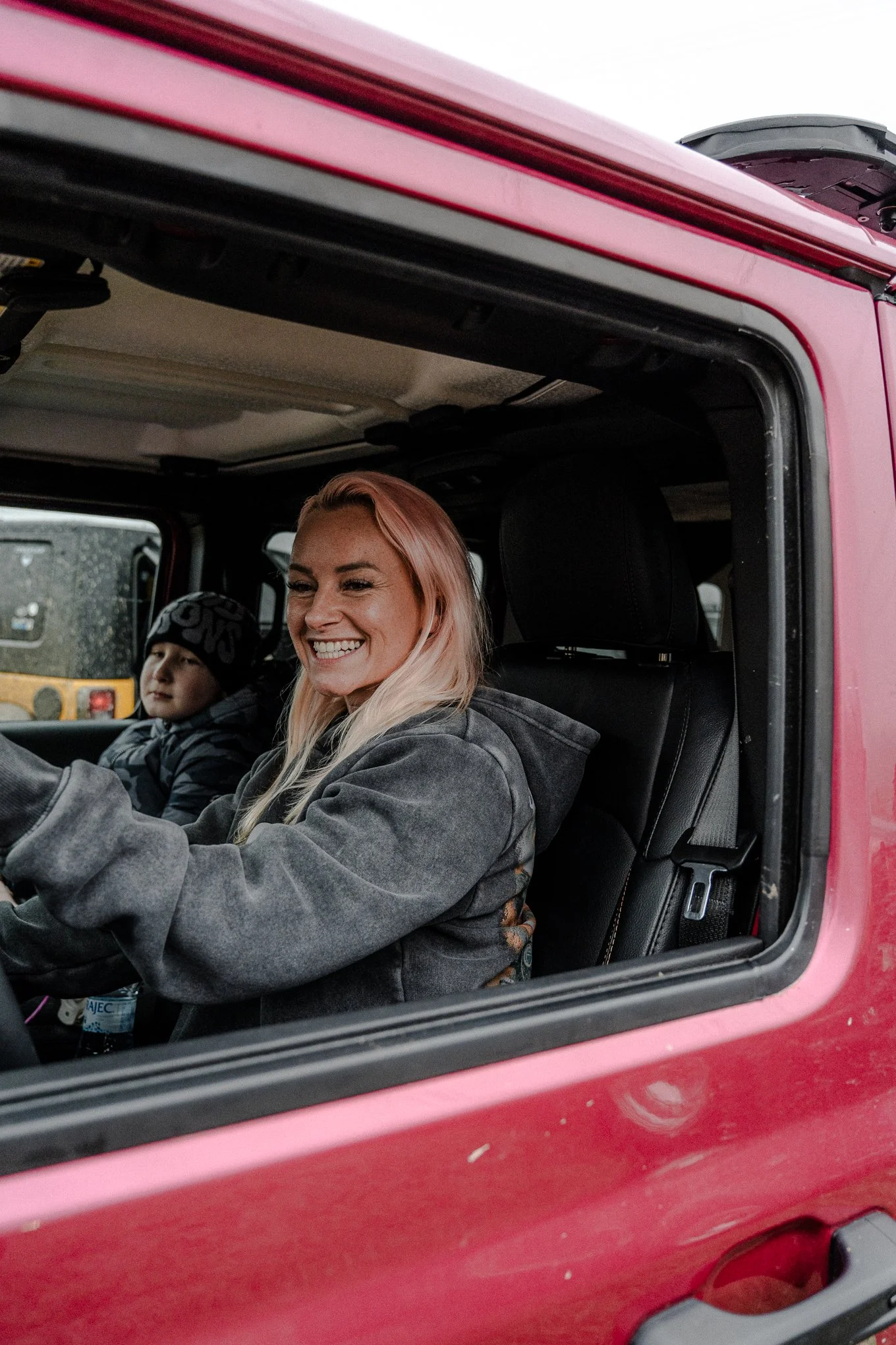 A woman with pink hair smiling inside a pink vehicle, sitting in the driver’s seat next to a young boy in the passenger seat, both wearing jackets.