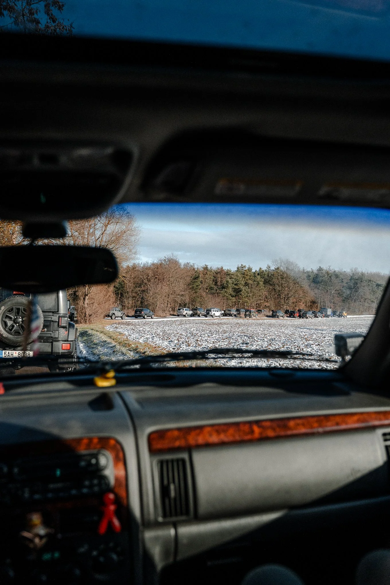 View from inside a car looking through the windshield at a line of parked vehicles on a snowy field with a forest in the background.