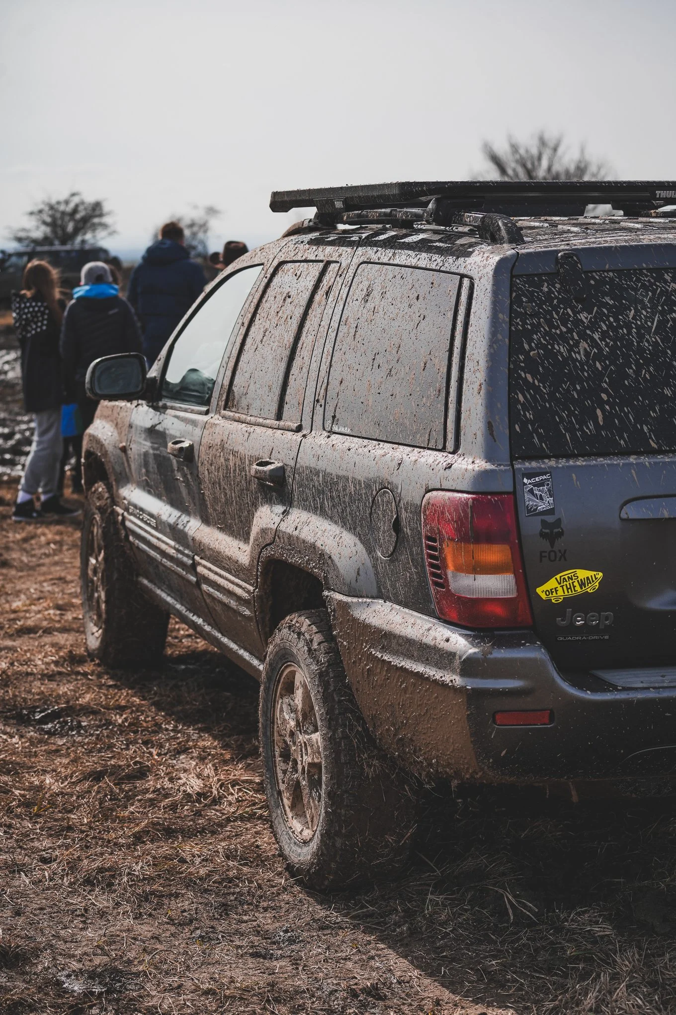 A muddy black SUV with stickers on the back, parked on a dirt field, with a group of people in the background.