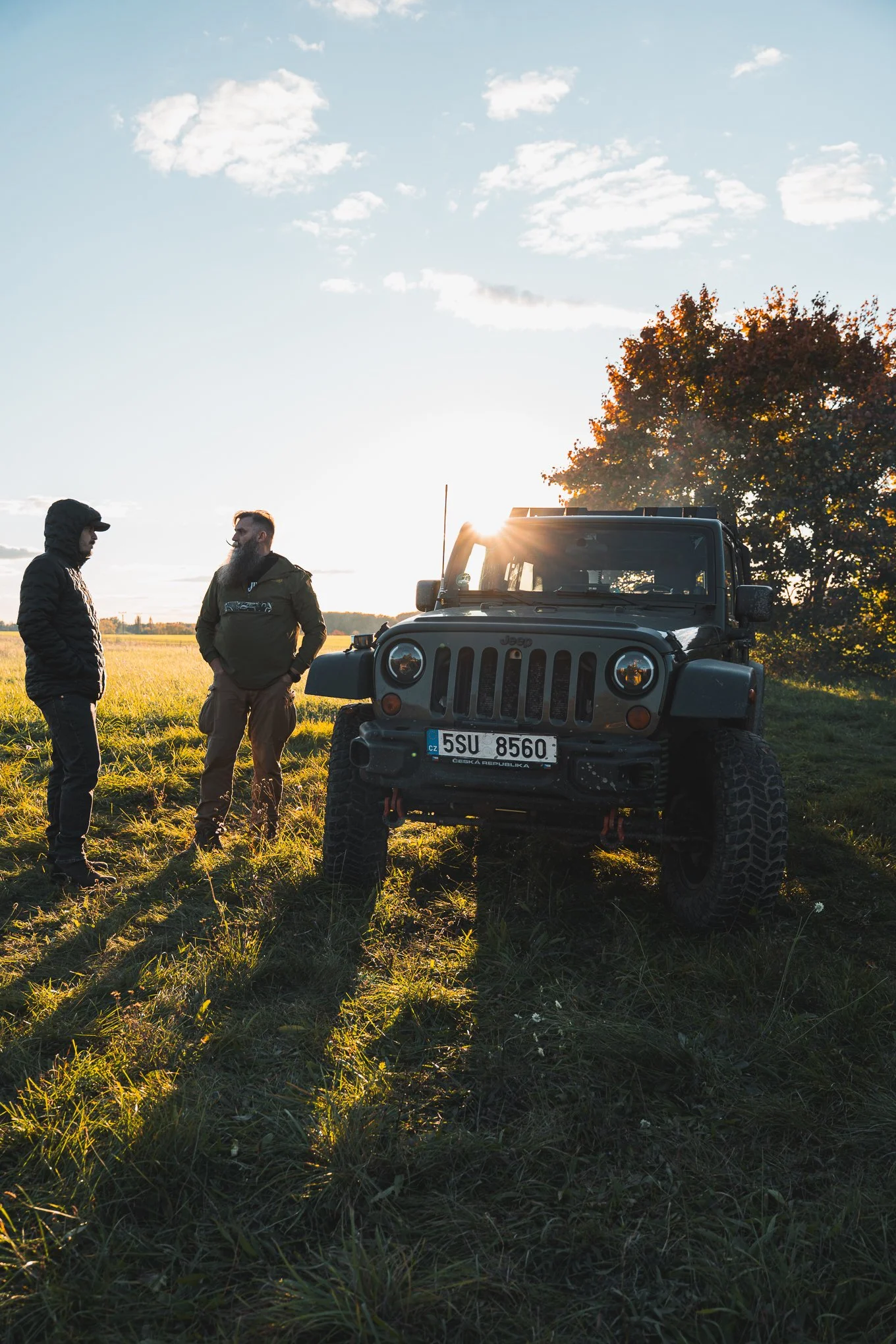Two men standing and talking next to a black Jeep in a grassy field at sunset with a tree and blue sky with clouds in the background.