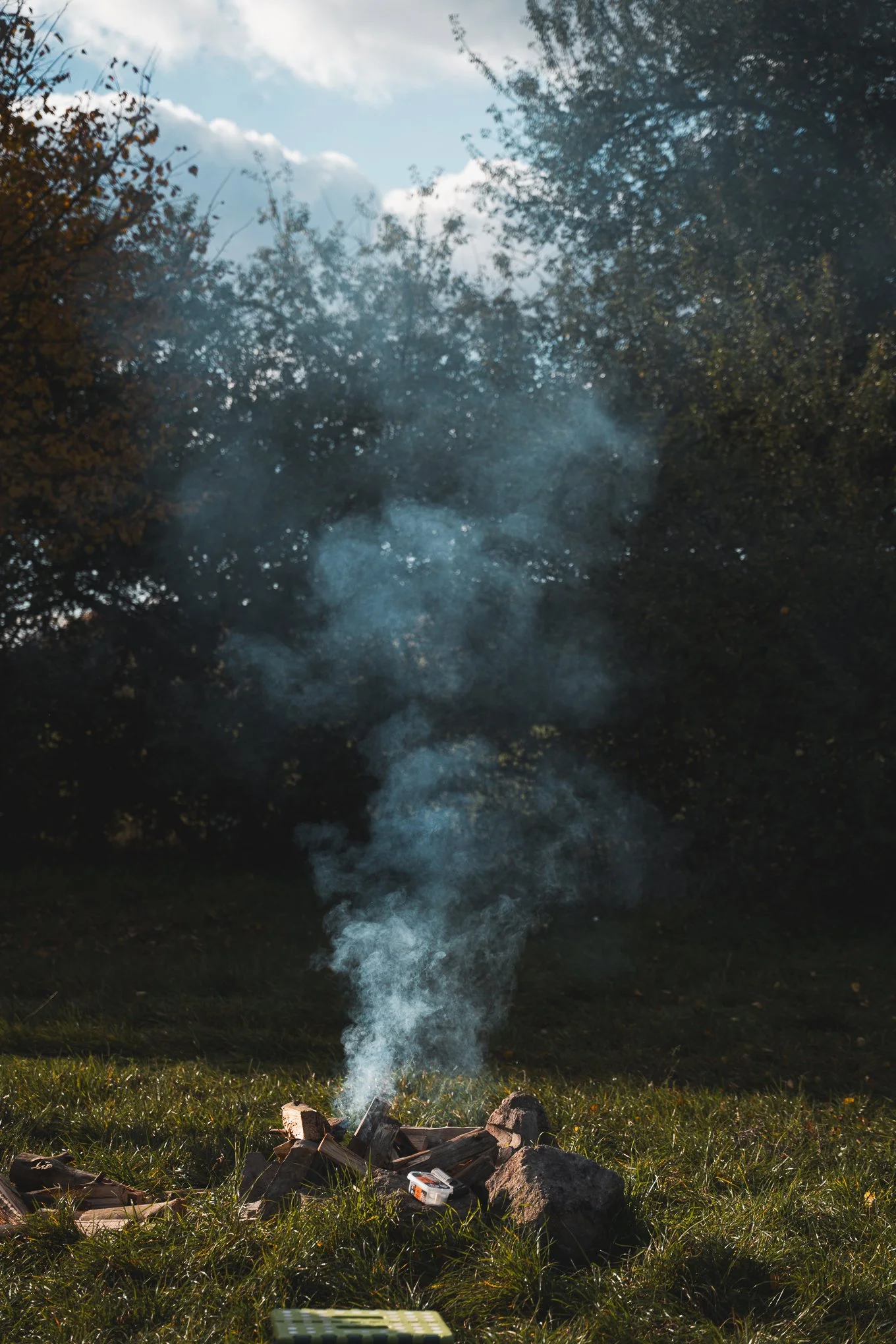 A small campfire surrounded by rocks with smoke rising into the sky in a grassy outdoor area, with trees and clouds visible in the background.