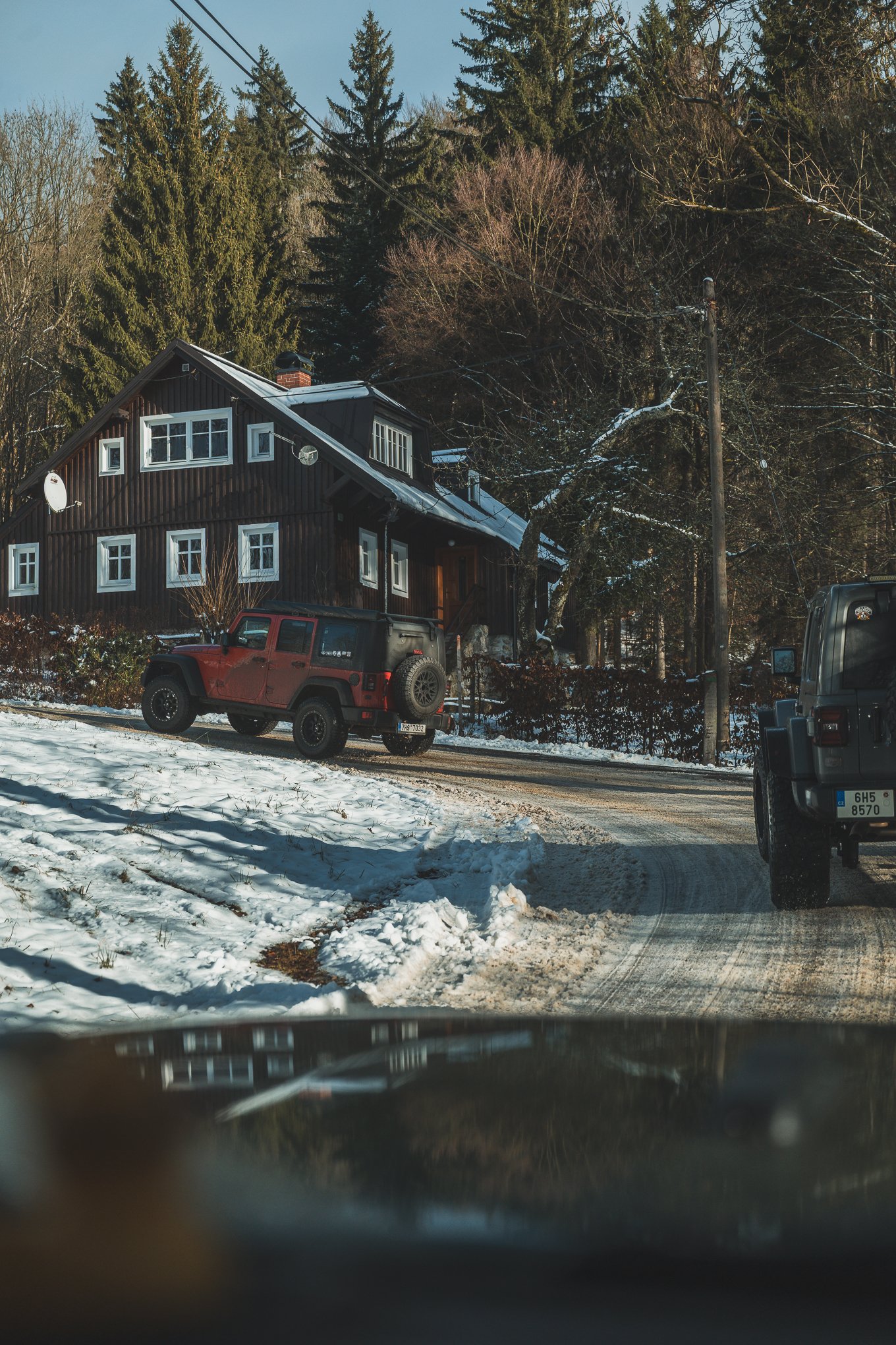 A snowy rural scene with a dark wooden house surrounded by tall pine trees. Two off-road vehicles are parked on a snow-covered and muddy driveway. Power lines run across the sky, and the sky is clear and blue.