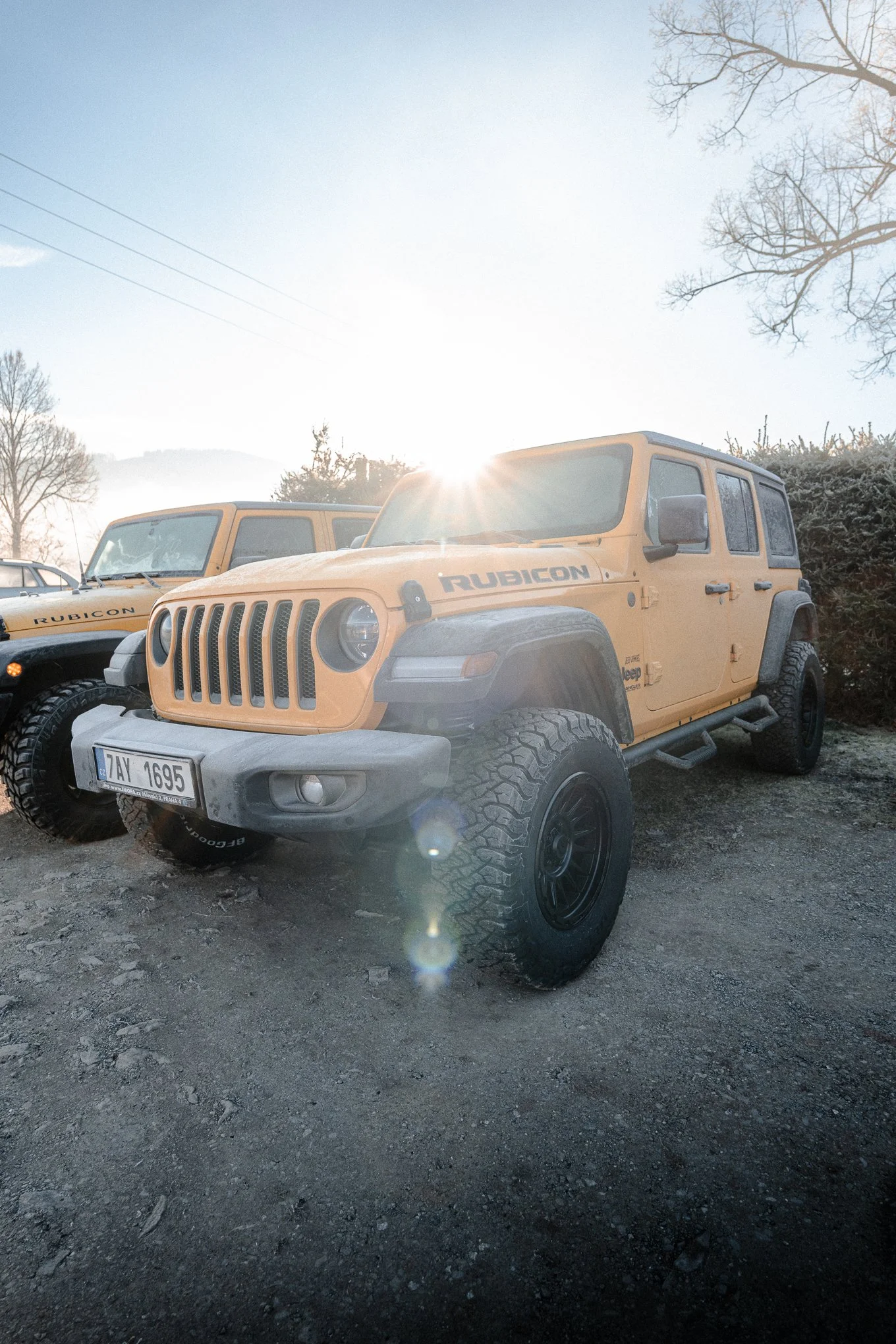 Yellow Jeep Rubicon parked on a dirt surface with another Jeep in the background, sunlight shining behind the vehicles, leafless trees in the background, clear sky.