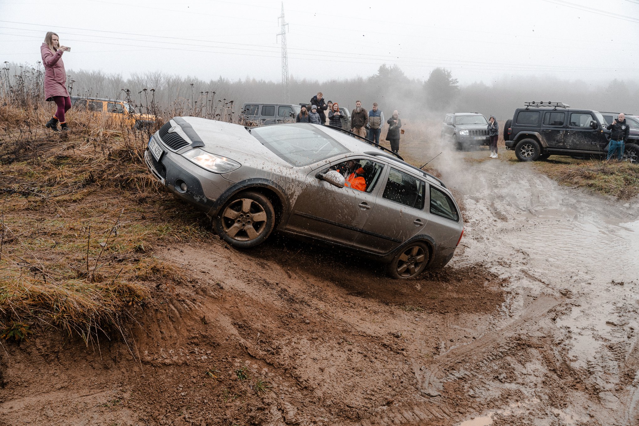 A silver car sliding down a muddy hill with the front wheels off the ground, surrounded by spectators and other vehicles on a foggy day.