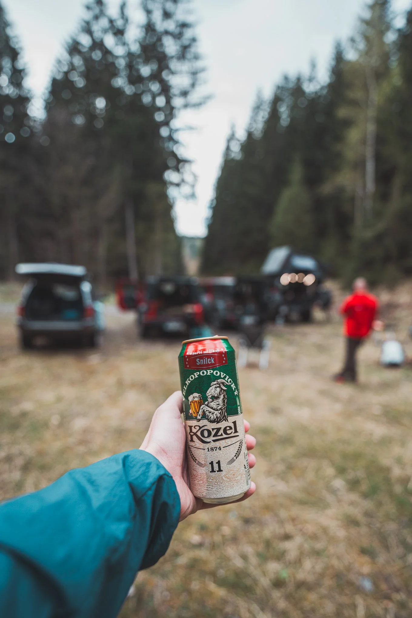 Person holding a can of Koze (Kozel) beer in front of a campsite with trees, cars, and two people in the background.