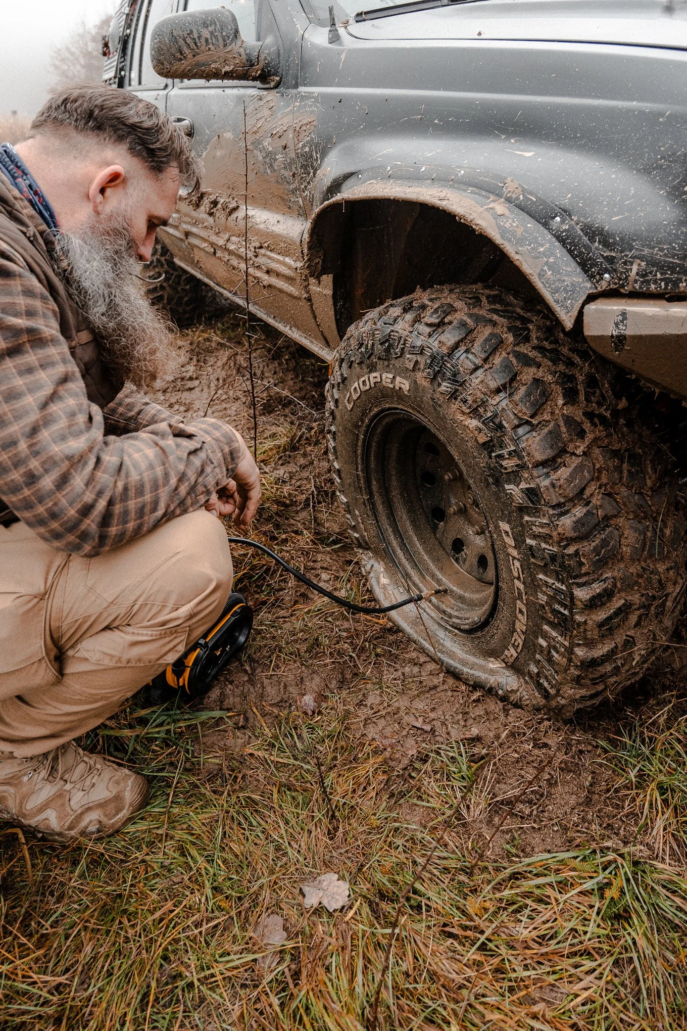 A man kneeling beside a muddy off-road vehicle, checking or adjusting a tire that is stuck in mud on a trail.