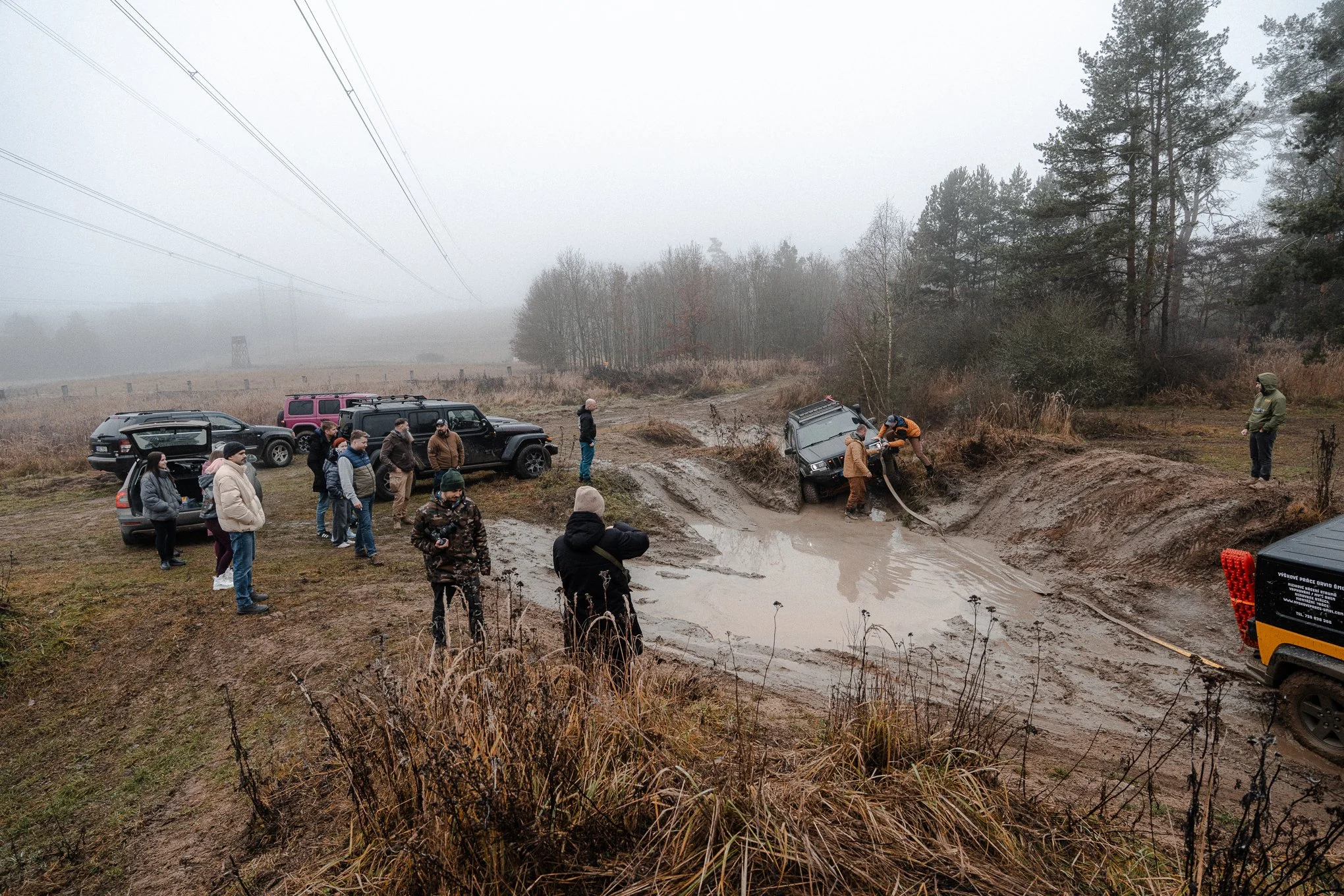 Group of people watching off-road vehicle rescue in muddy water-filled ditch on foggy day with trees and power lines in background.