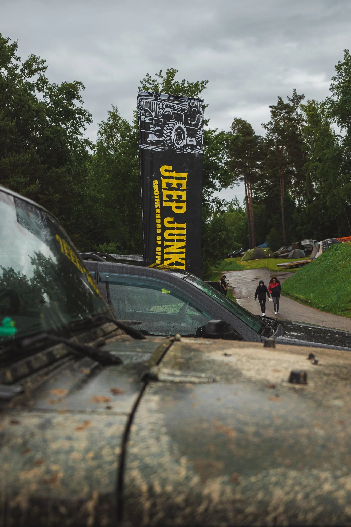A cloudy outdoor scene with a Jeep parked in the foreground, a tall Jeep JUNKY banner with a graphic of a Jeep vehicle, and a dirt trail with tents and two women walking in the distance surrounded by trees.