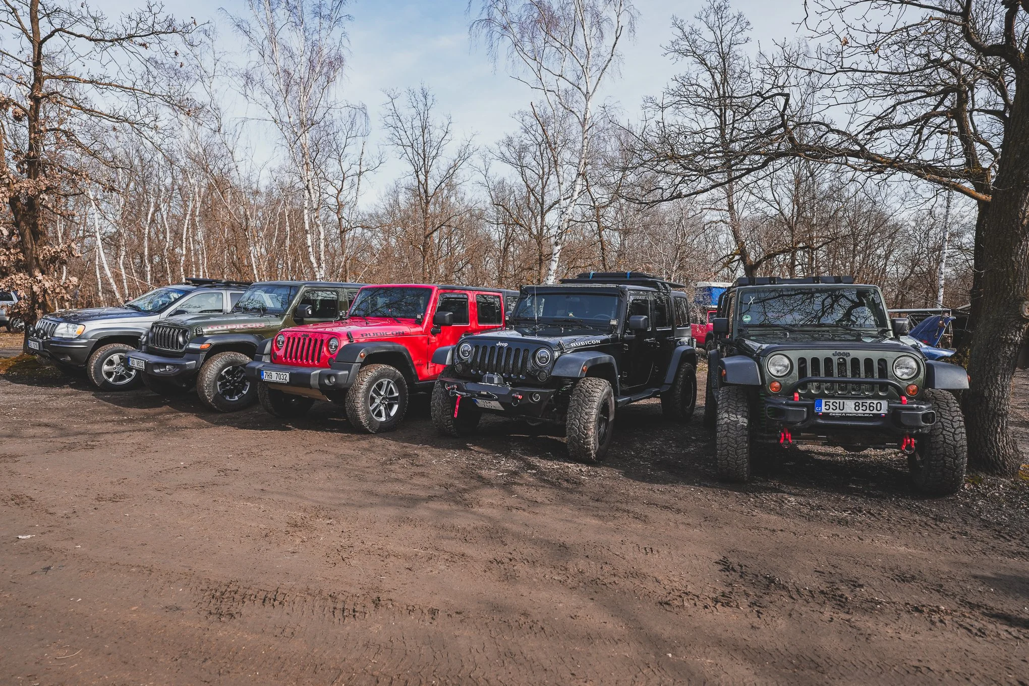 A lineup of various Jeep vehicles parked outdoors on dirt ground with leafless trees in the background, including two black Jeeps, a red Jeep, and a silver Jeep.