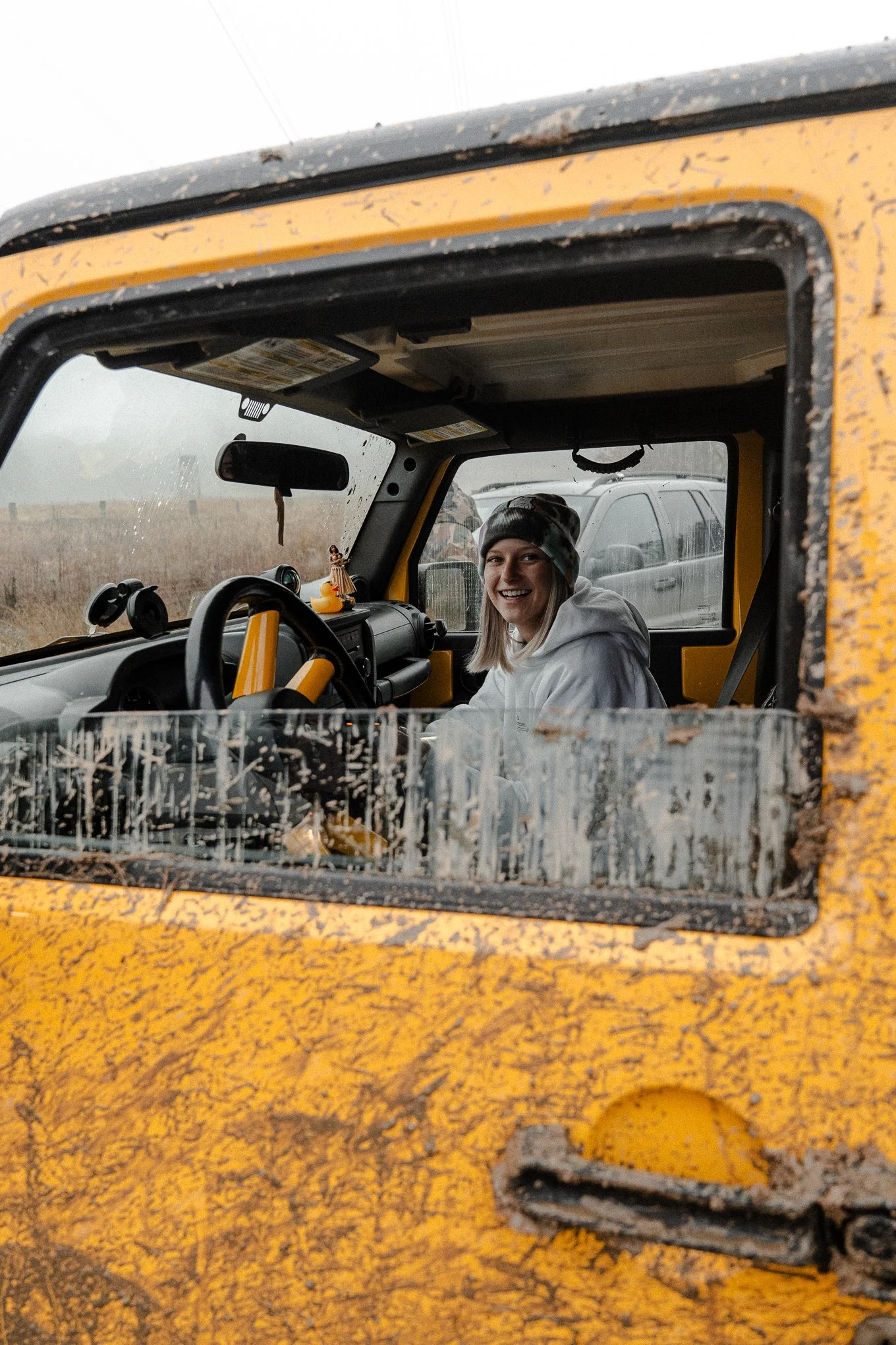 A woman smiling and sitting inside a yellow, muddy off-road vehicle, wearing a grey hoodie and a camo beanie, in an outdoor rural setting.