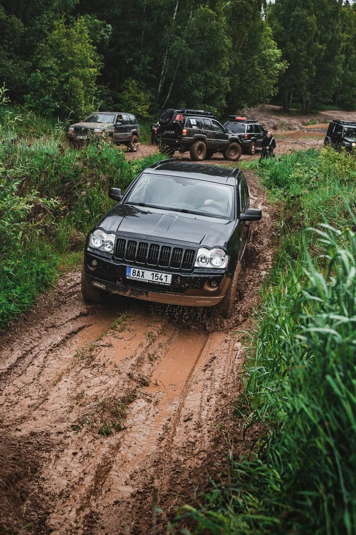 A black SUV driving through a muddy off-road trail surrounded by green grass and trees, with several other vehicles parked in the background.