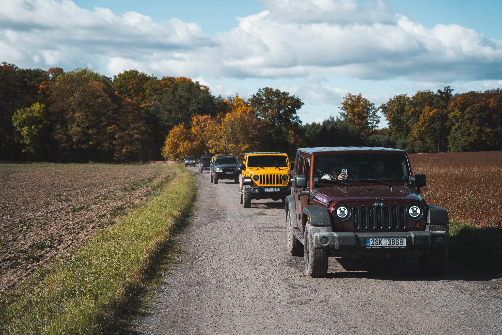 A line of five Jeep vehicles, including a maroon Jeep in the front, traveling on a gravel country road surrounded by fields of crops and trees with autumn foliage under a partly cloudy sky.