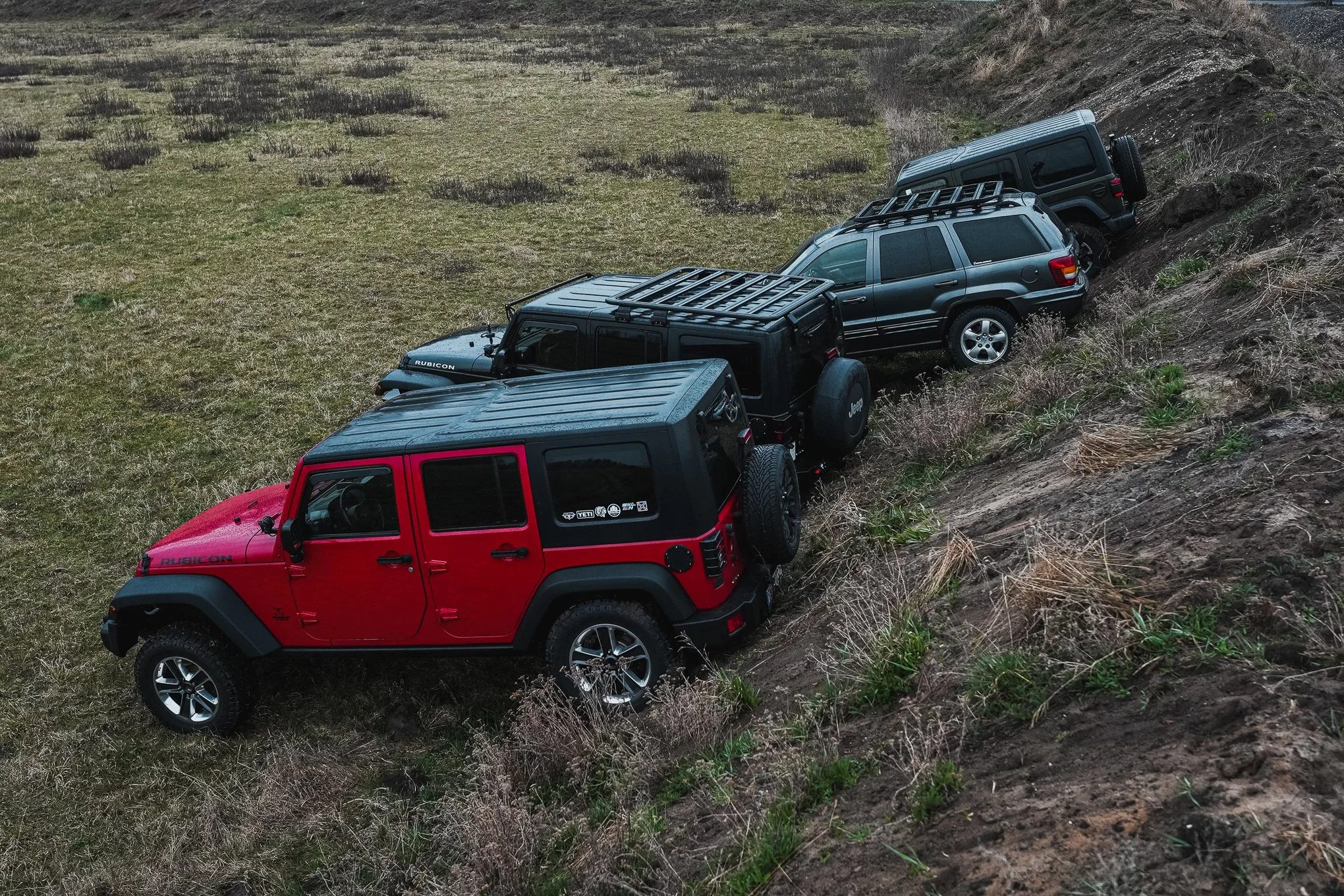 Four off-road vehicles parked on a grassy slope, with the red Jeep at the front and the others in a line behind it, ascending the hill.
