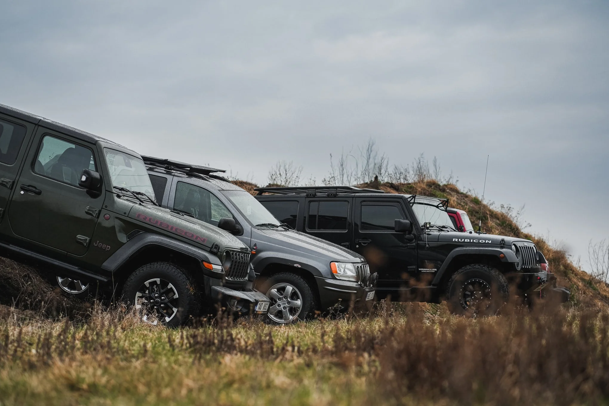 Three Jeep vehicles parked on a grassy hillside under a cloudy sky, with the first vehicle being a black Jeep Rubicon, followed by a gray Jeep and a dark gray Jeep, all equipped for off-road driving.