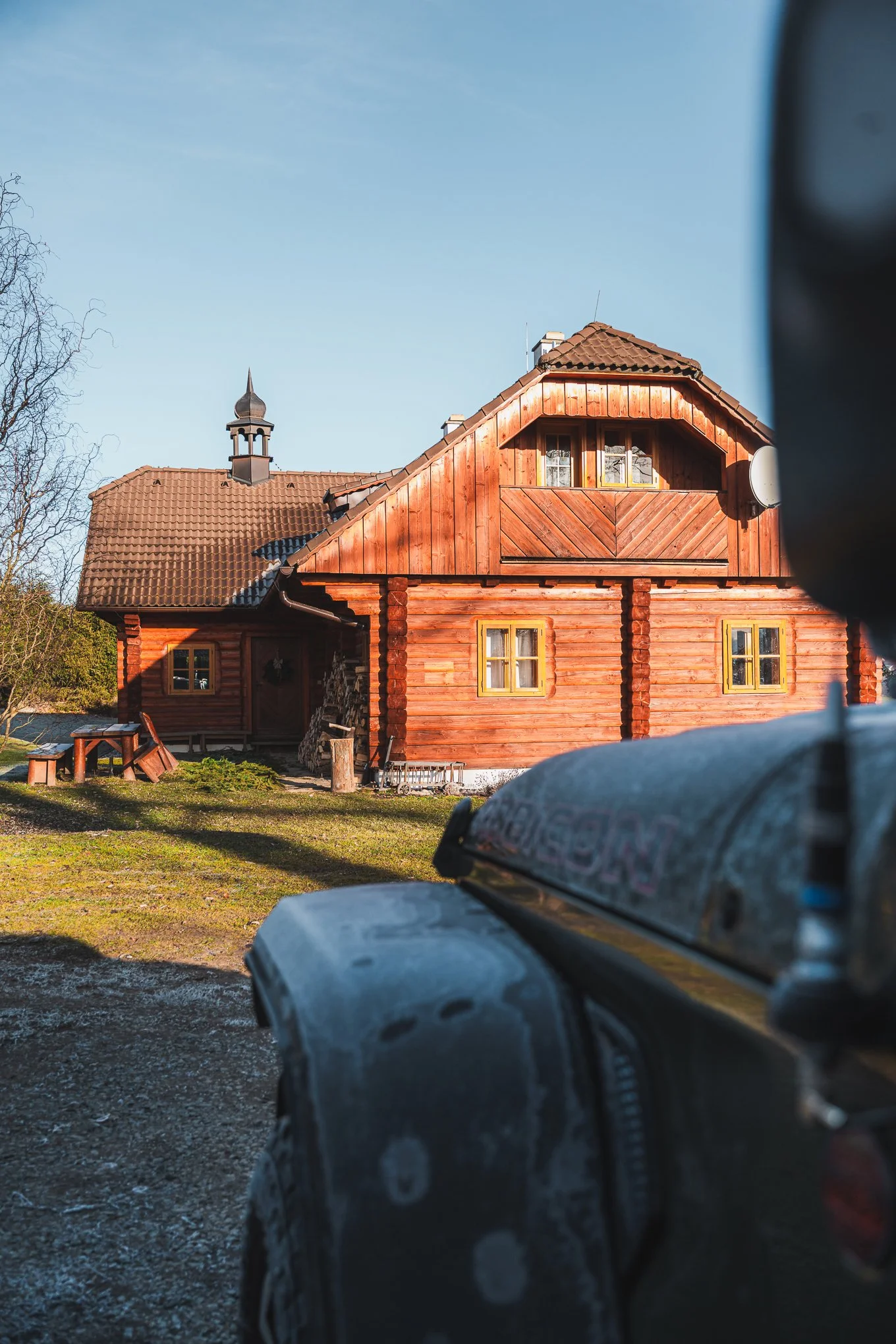A wooden house with a red-brown exterior, multiple windows with yellow trim, a small balcony, and a steeply pitched roof with a small tower, set against a clear blue sky. The foreground shows part of a dark vehicle.