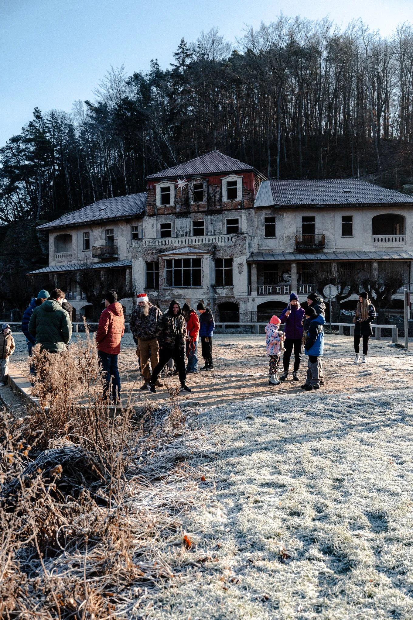 A group of people, including children and adults, gathered outside on a frosty day in front of a large, old, abandoned house surrounded by trees.