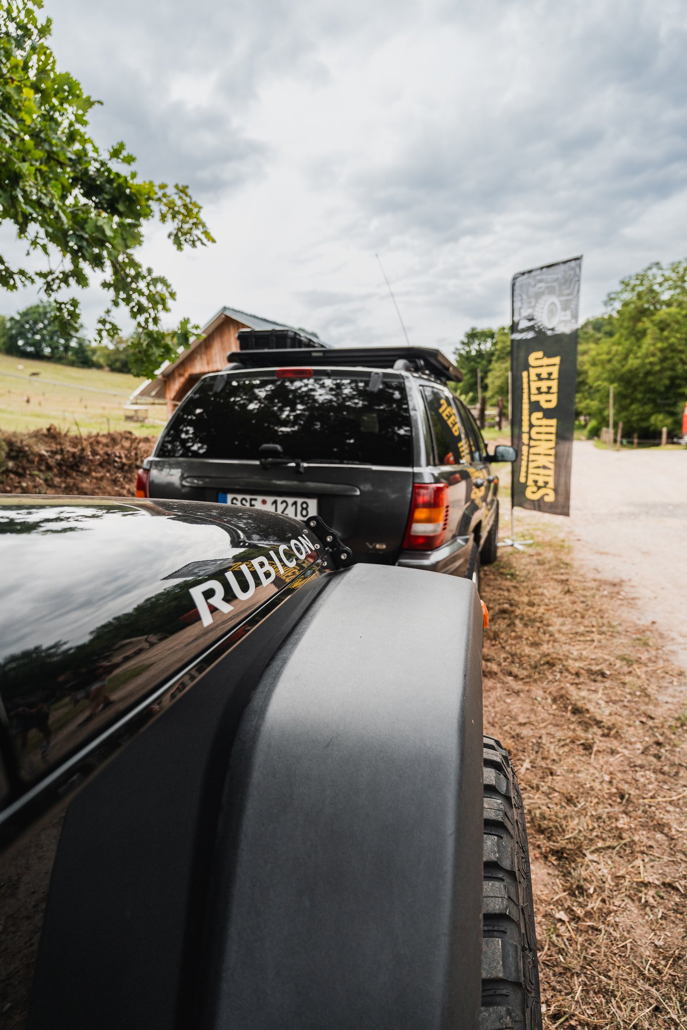 Close-up of a black vehicle with the word 'RUBICON' on the side mirror, parked next to a gray vehicle on a dirt path with trees and a banner in the background.