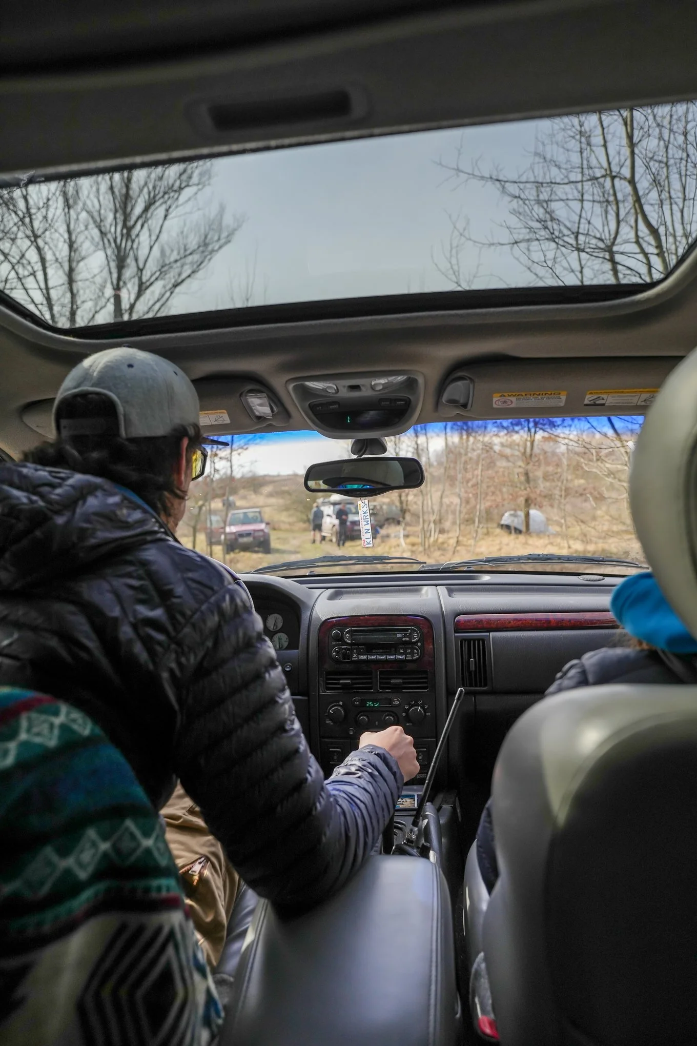 View from inside a vehicle, showing two people in the front seats, with a man on the left adjusting the gear shift and a woman on the right. Outside, there are trees and a few parked cars on a cloudy day.