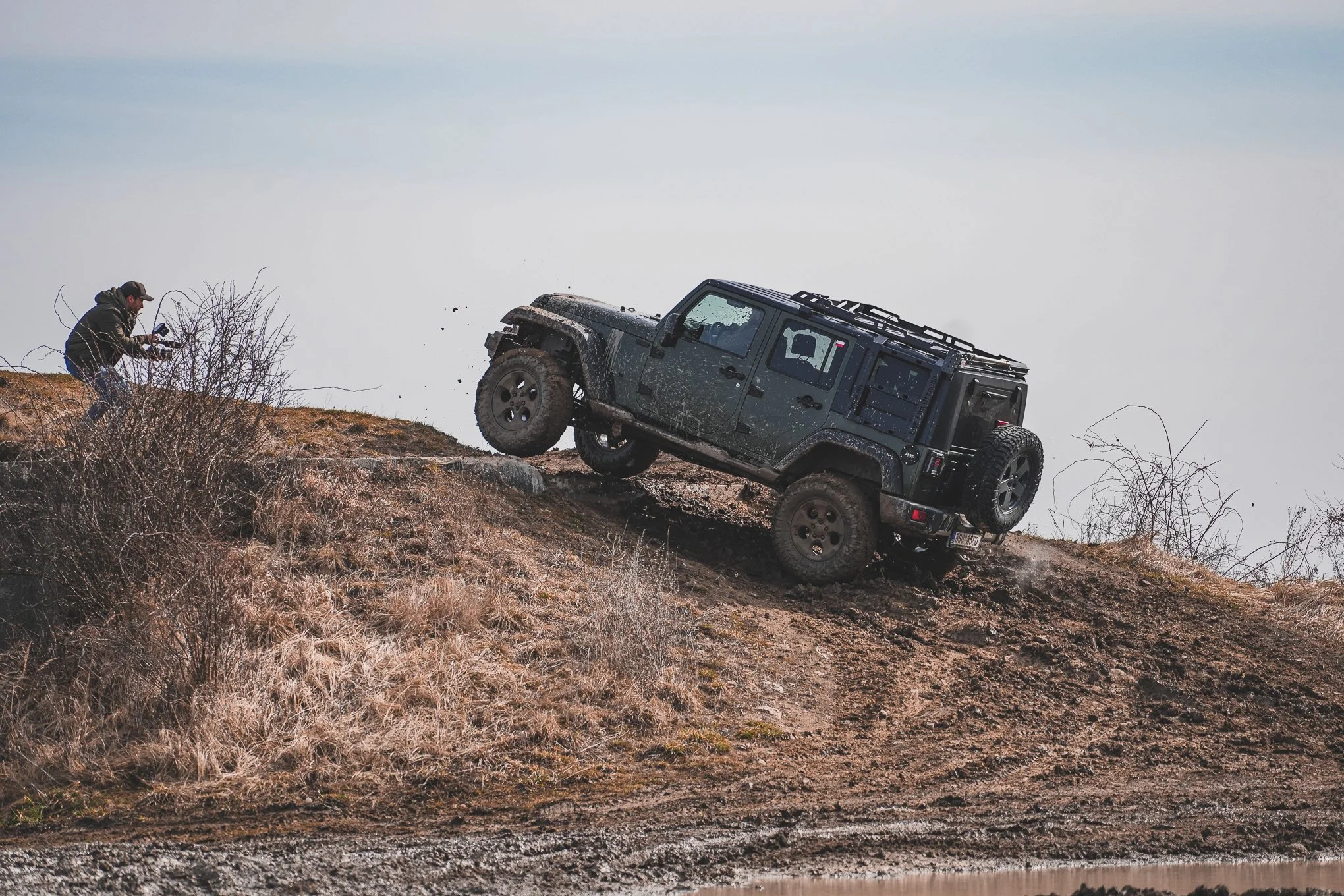A black off-road vehicle climbing a steep hill with mud on its tires, while a person takes a photo or records video nearby.