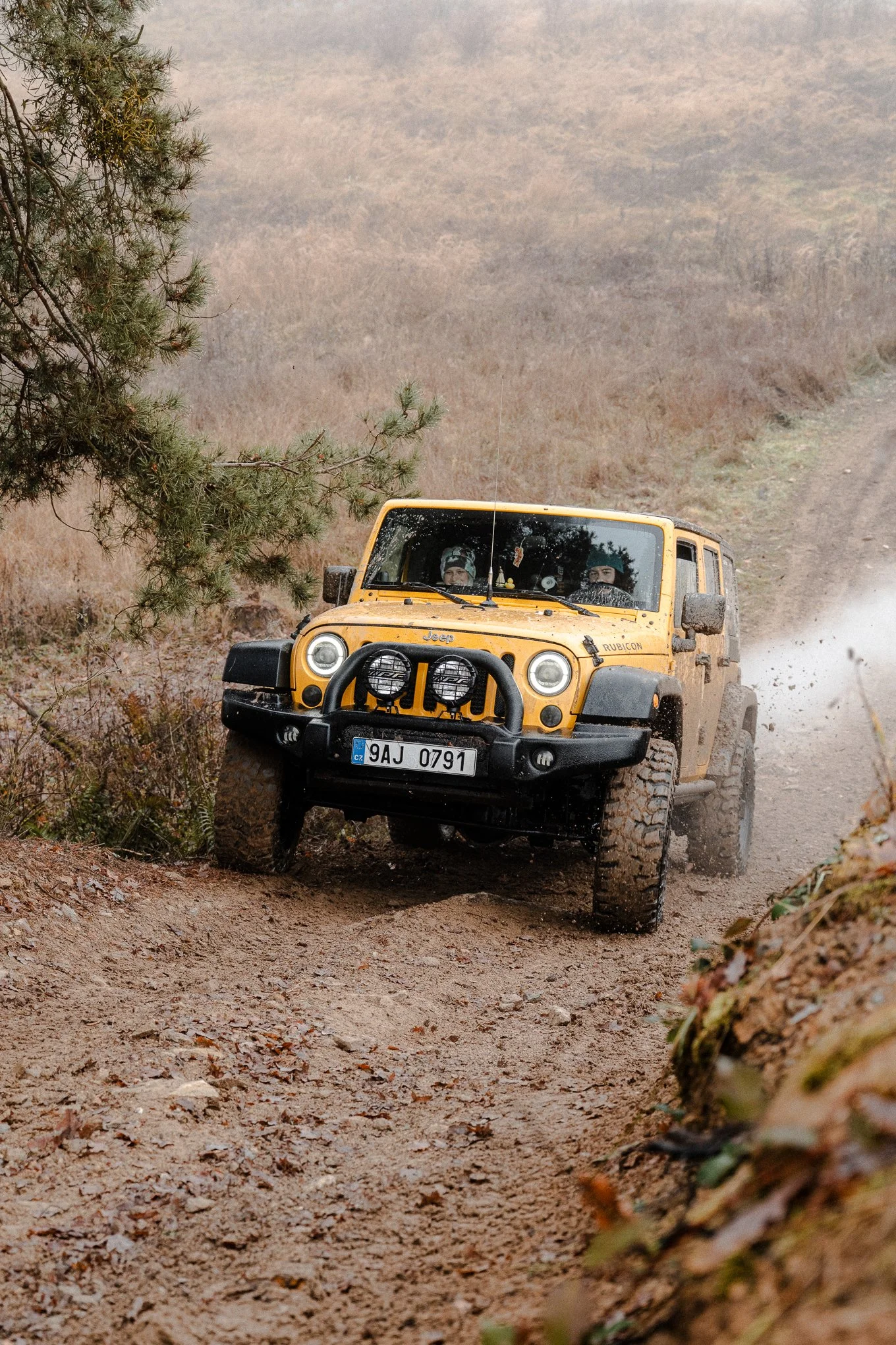A yellow Jeep Rubicon driving on a muddy off-road trail in a mountainous area, kicking up dirt with two drivers visible through the windshield.