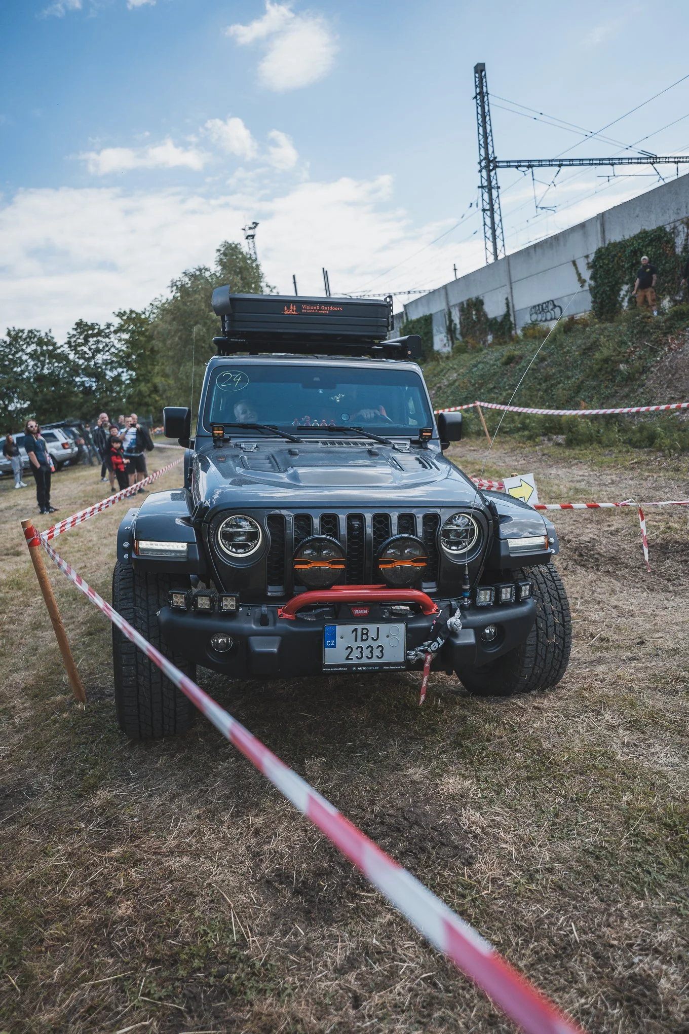A black Jeep Wrangler with off-road lights and equipment, parked on a grassy area at an outdoor event, with people in the background and a cloudy sky overhead.