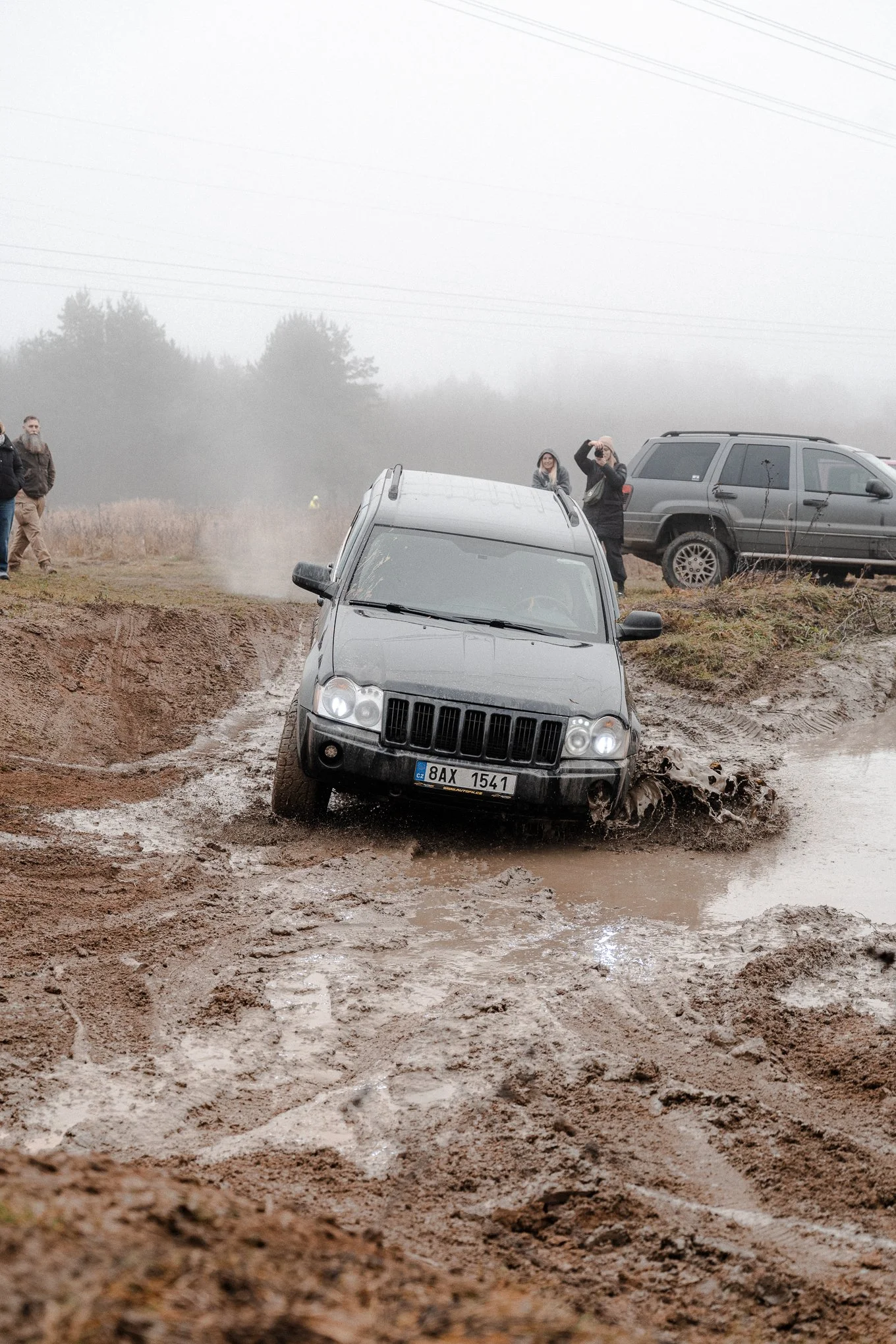 A black SUV driving through a muddy off-road trail with muddy water splashing around its tires, while a group of people observe nearby on a foggy day.