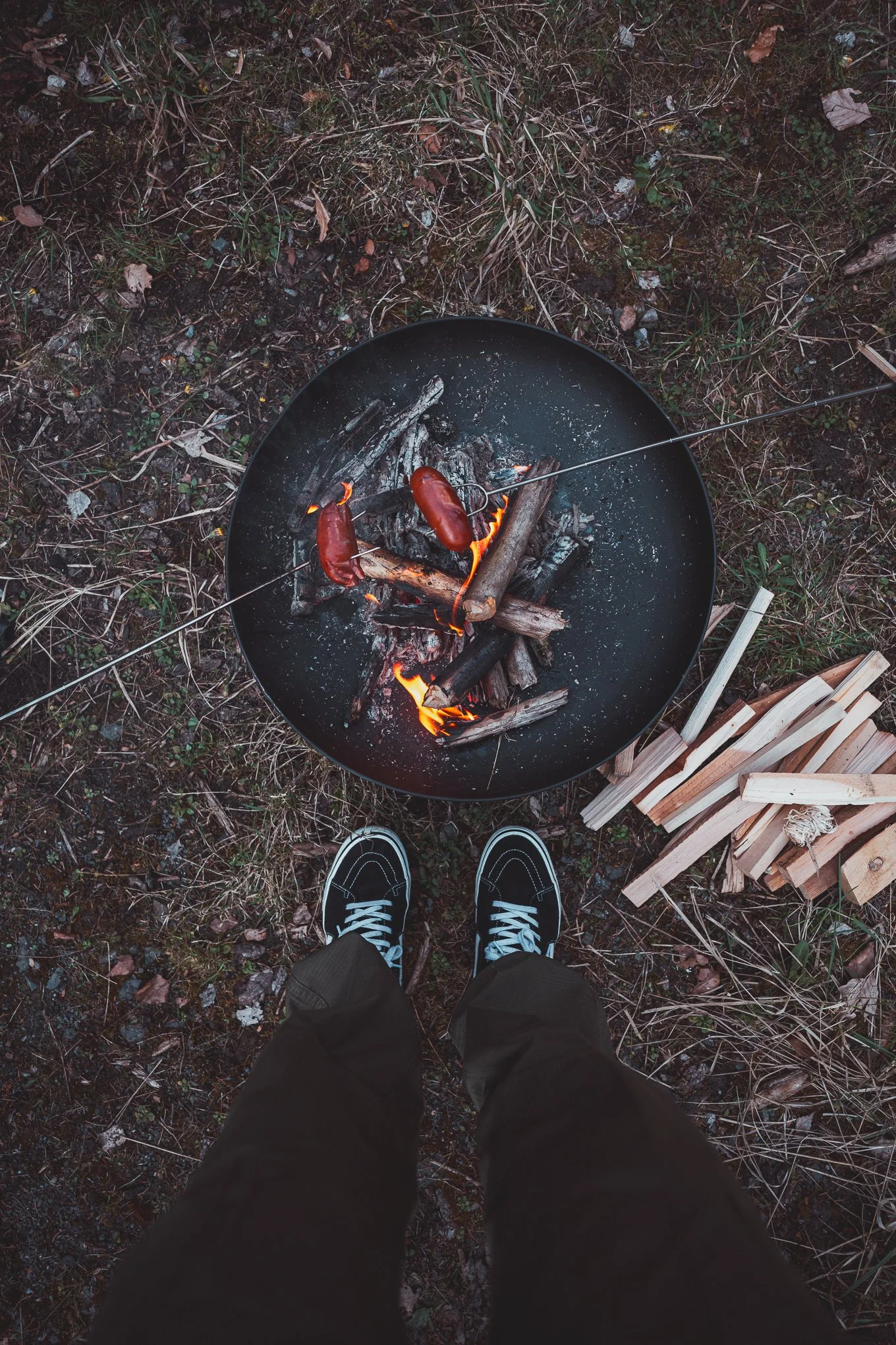 A person standing outdoors wearing black shoes and pants, looking down at a fire pit with burning wood and two sausages on skewers, and a small pile of chopped wood nearby.
