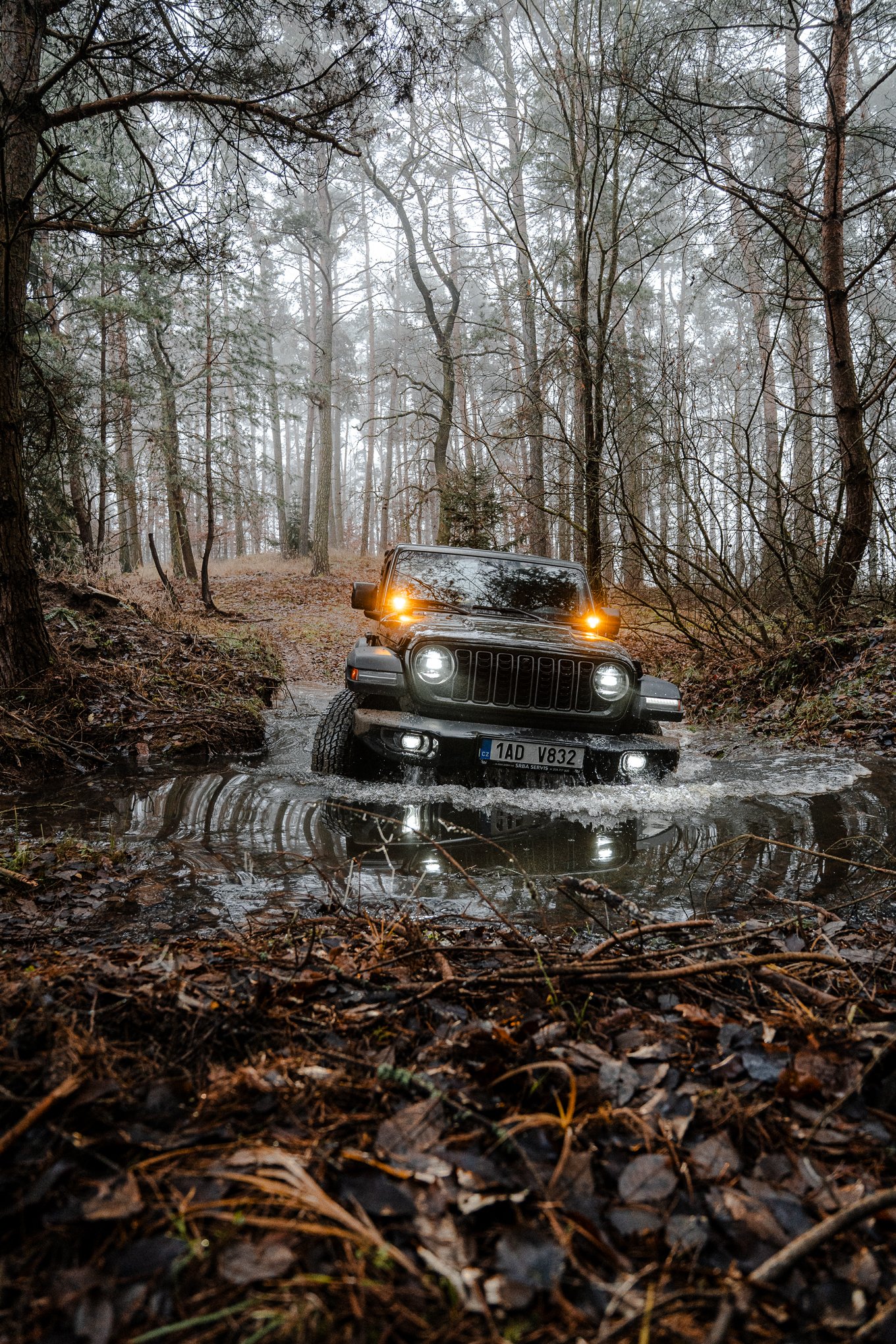 A black Jeep vehicle driving through a water crossing in a forest, with foggy weather and leaf-covered ground.