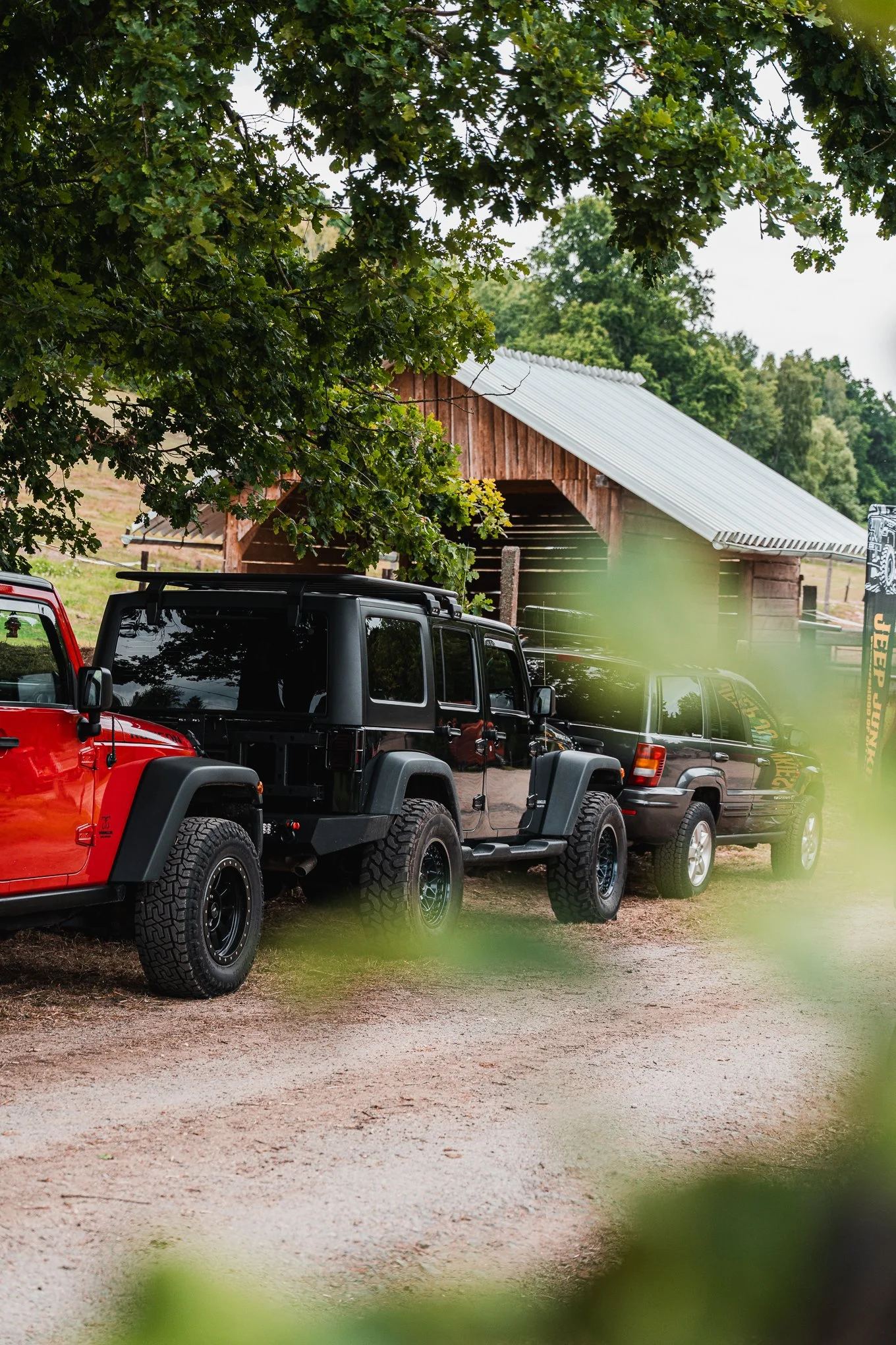 A row of parked off-road vehicles, including a red Jeep and a black Jeep, in front of a wooden barn on a farm with green trees in the background.