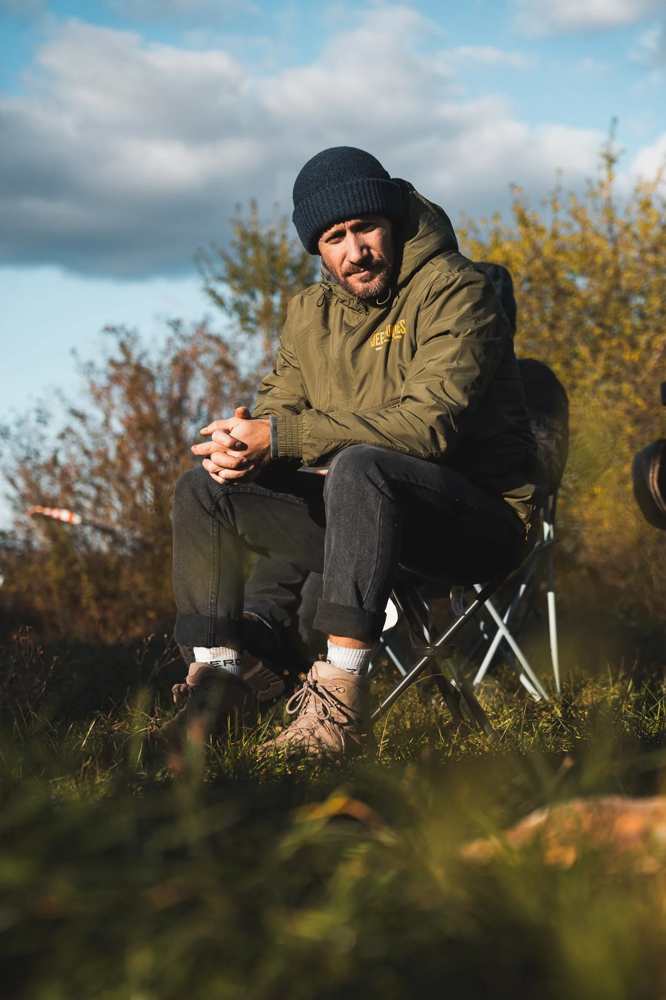 Man sitting on a portable camping chair outdoors during daytime, wearing a beanie and a green jacket, with trees and clouds in the background.