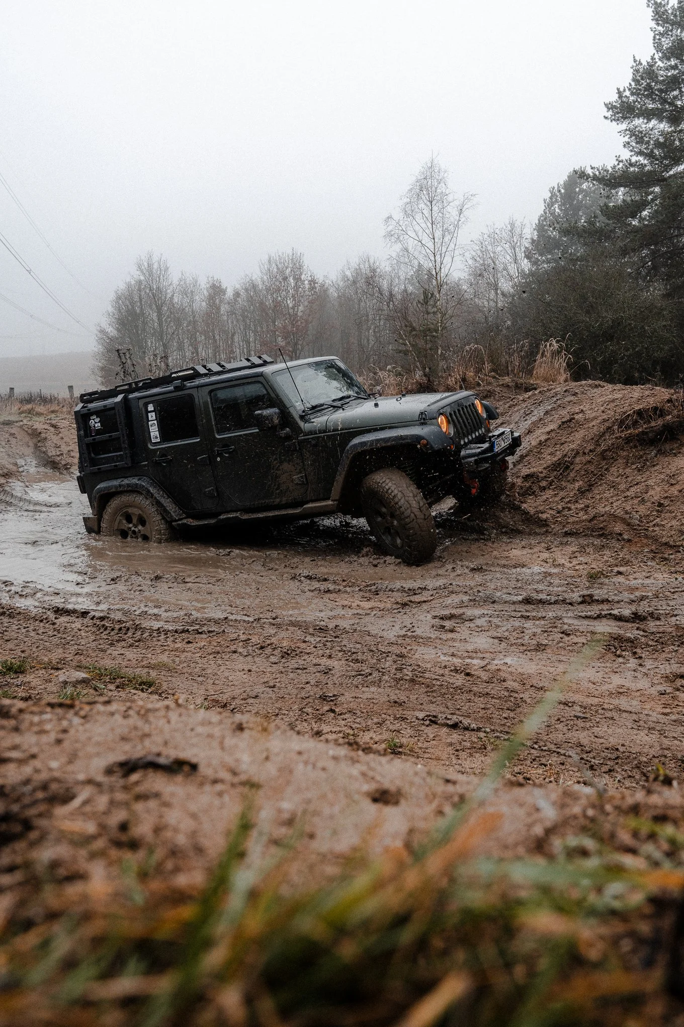 A black Jeep off-road vehicle with muddy wheels partially stuck in a muddy trail with trees and gray sky in the background.