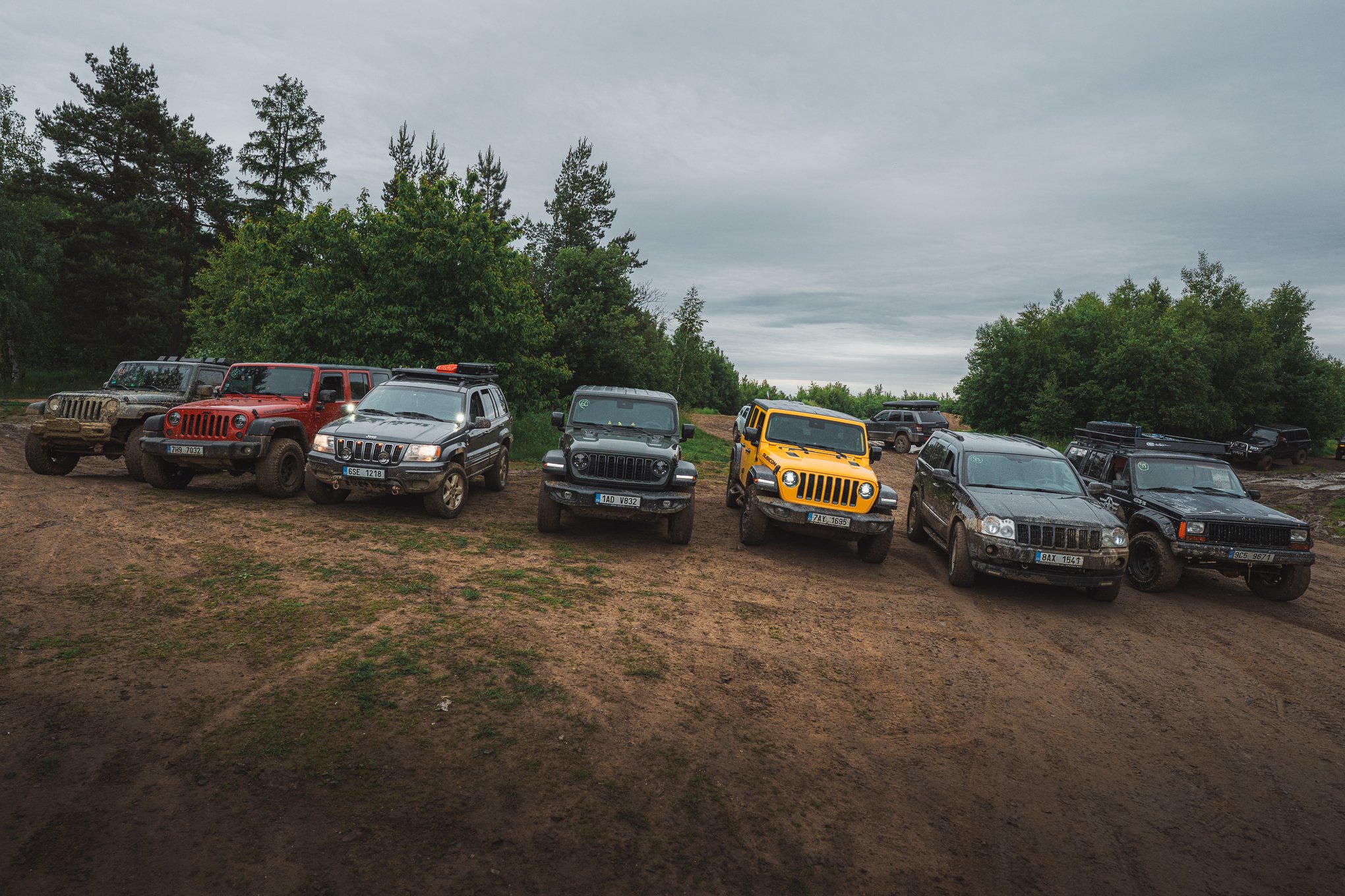 A lineup of seven off-road vehicles, including Jeeps and SUVs, parked on a dirt hill in a forested area under a cloudy sky.