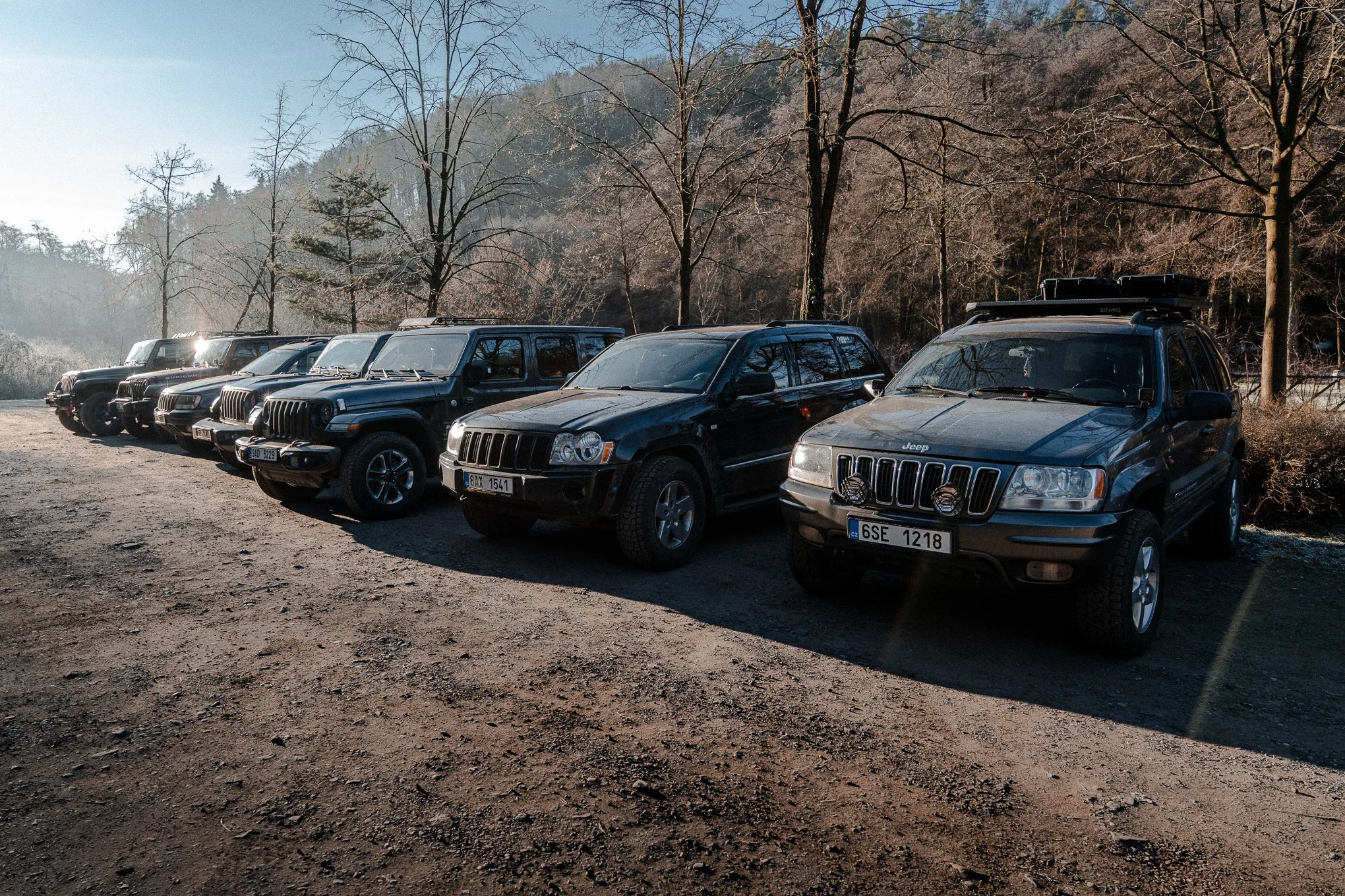 A row of six black and dark gray Jeep vehicles parked on a dirt surface, with trees and hills in the background.