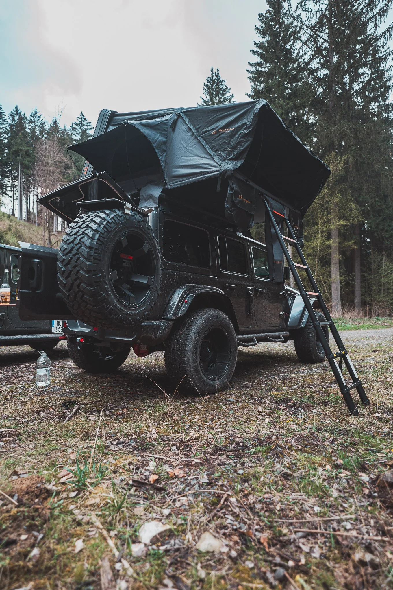 A black off-road vehicle with a rooftop tent set up and a ladder leaning against it, parked on a wooded area with trees in the background.