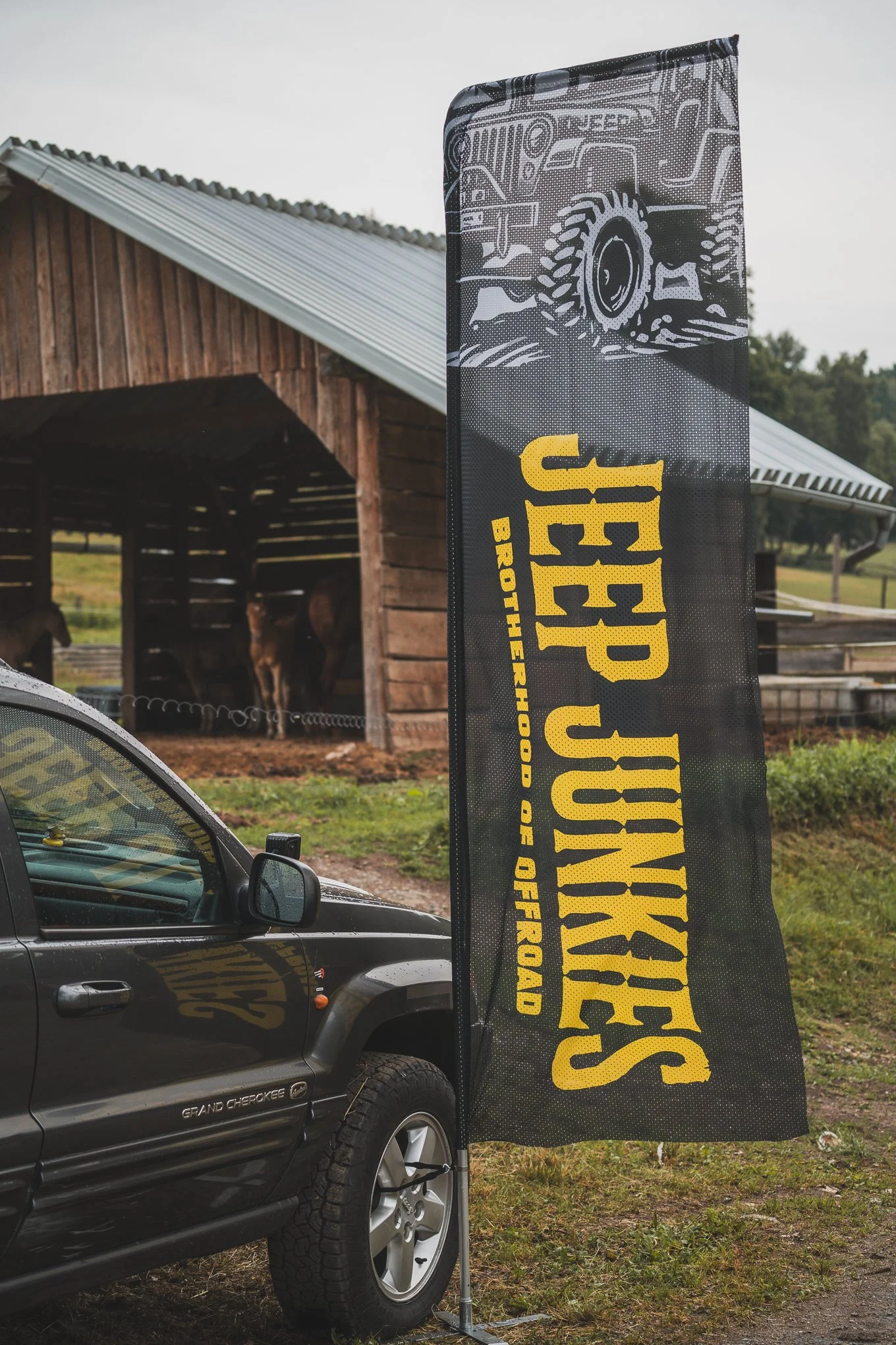 A black Jeep Grand Cherokee parked outdoors near a farm building with a metal roof. A tall black flag with yellow and white text and graphics is displayed next to the vehicle, promoting Jeep Jinkings Brothers Highway of Offroad.