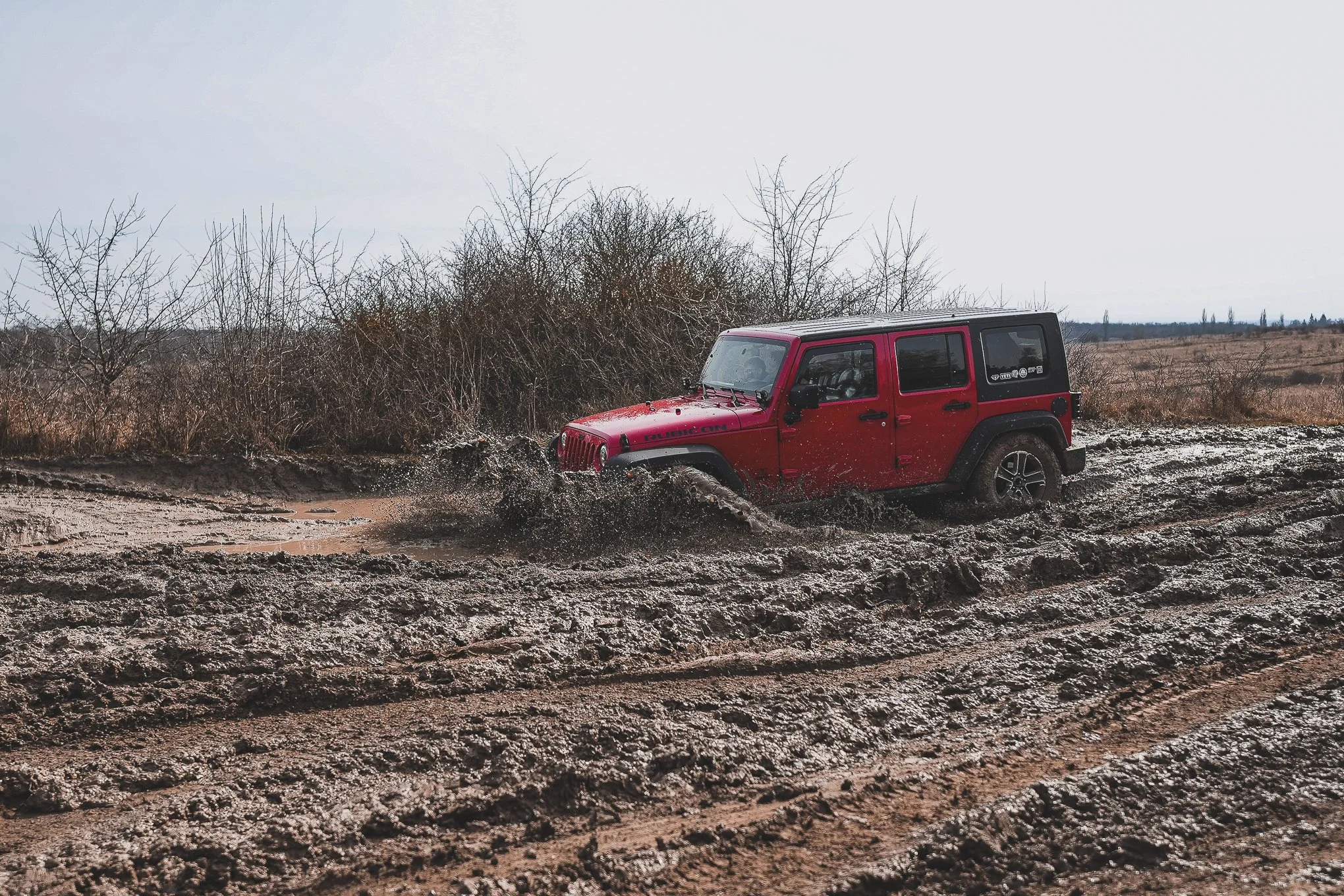 Red Jeep driving through muddy off-road terrain with bushes and open sky in the background.