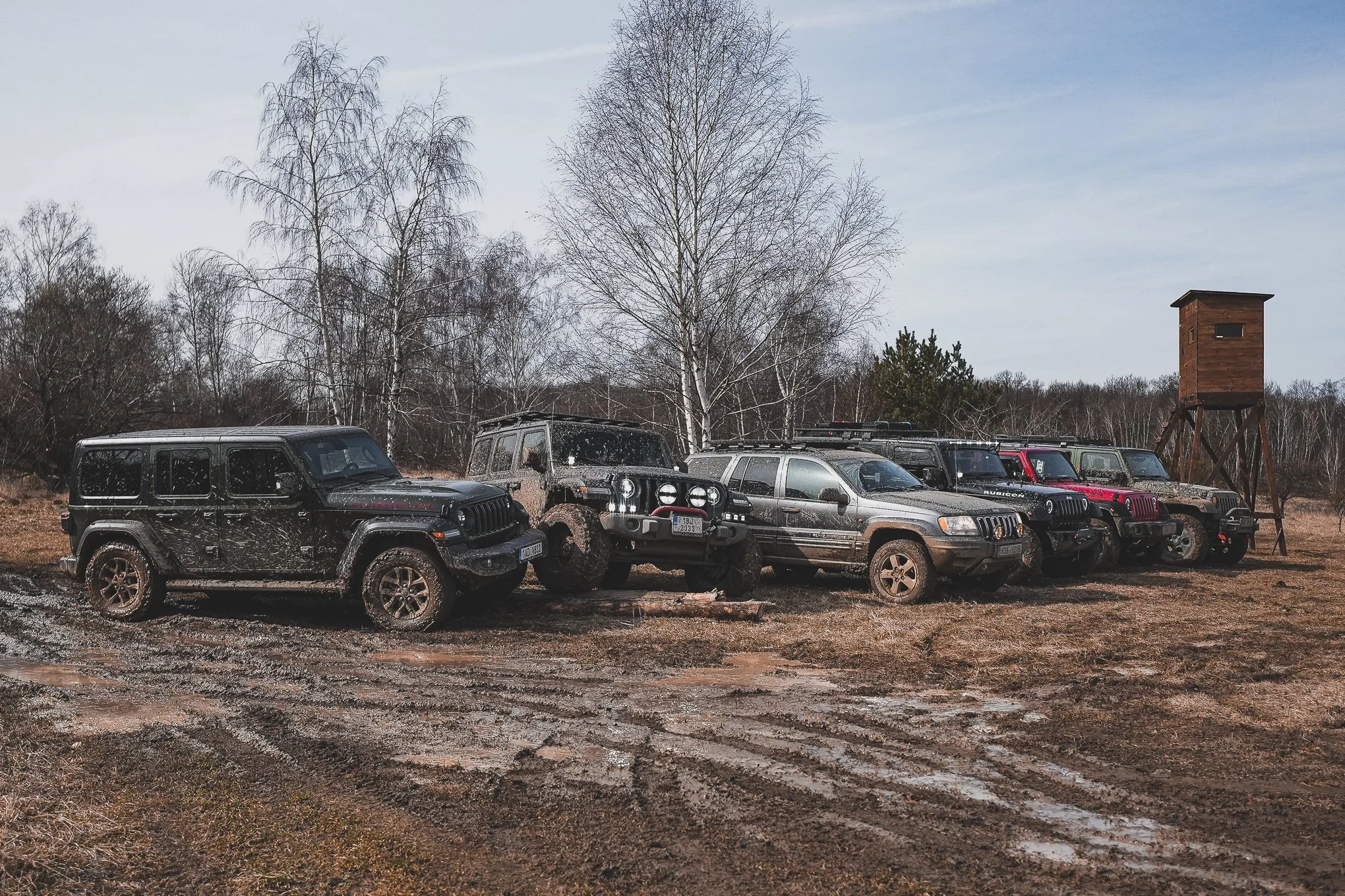Multiple off-road vehicles covered in mud parked in a muddy field near leafless trees and a wooden lookout tower.