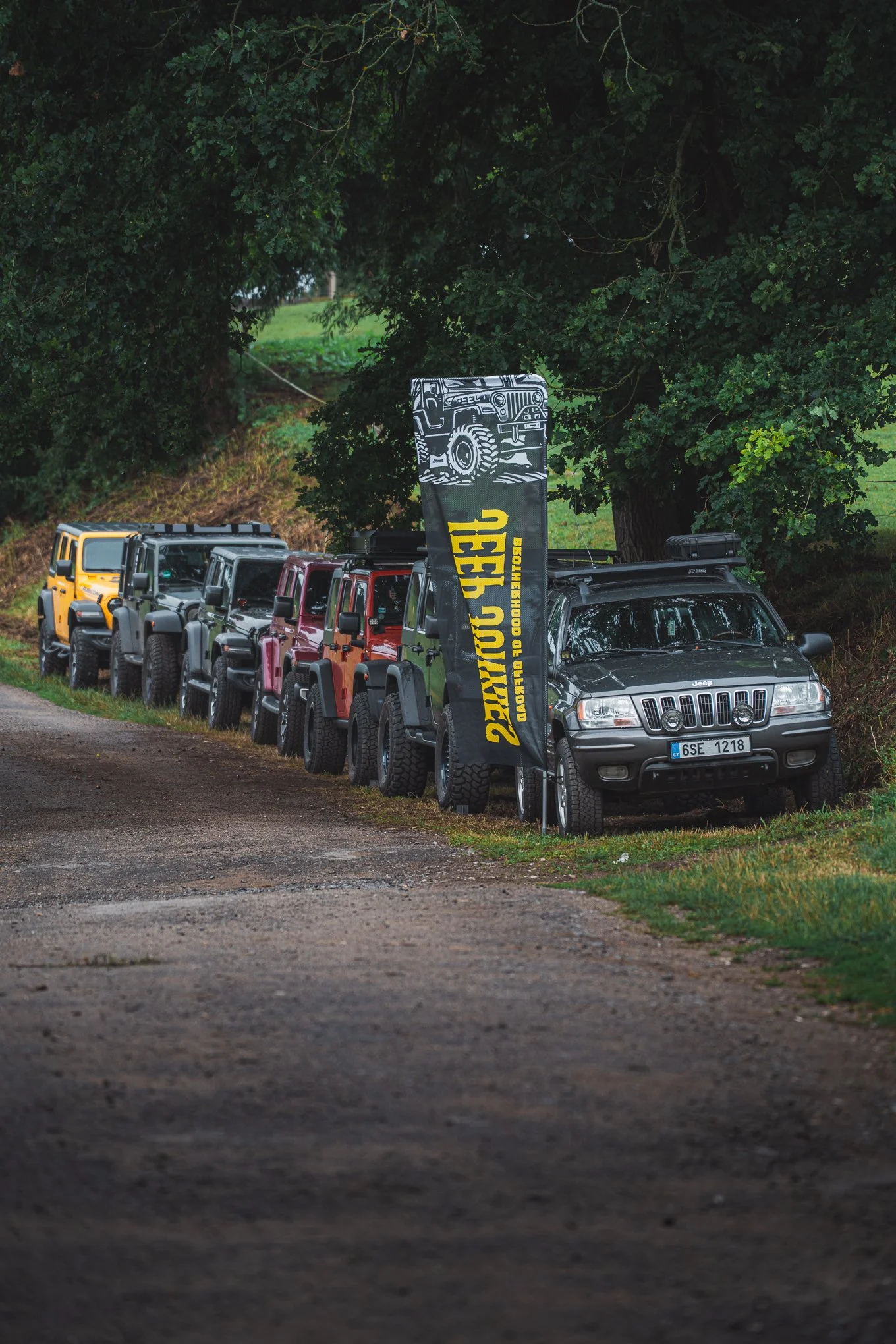 Multiple parked off-road vehicles in a row along a dirt path with trees and grass in the background, and a flag with a graphic of a vehicle and yellow text.