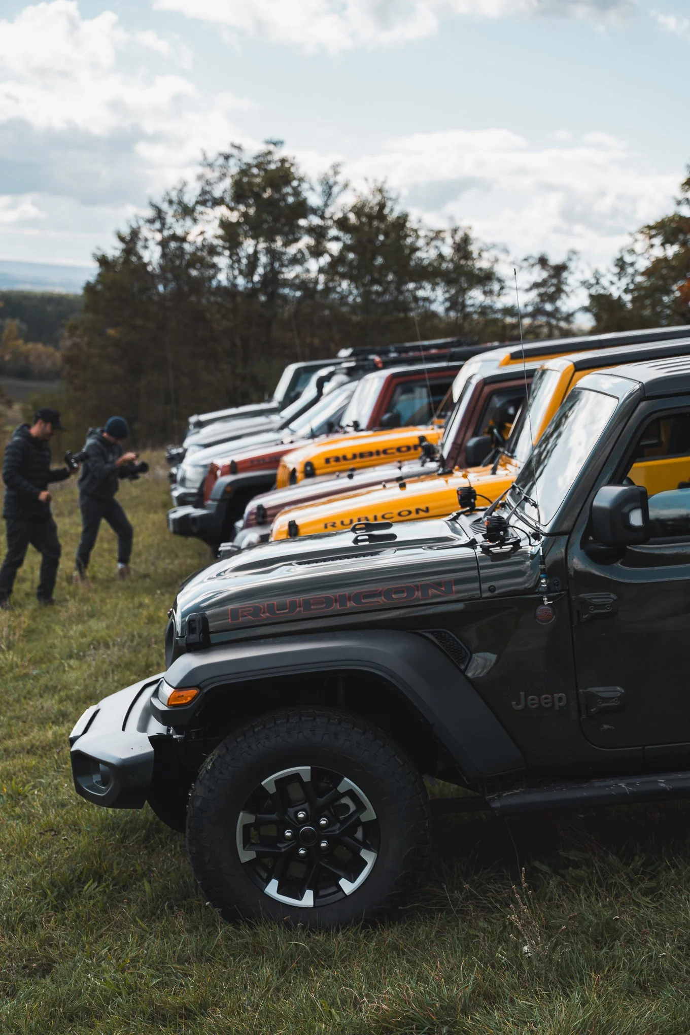A lineup of Jeep Rubicon vehicles parked on a grassy field, with two people taking photos or videos in the background, and trees and a cloudy sky in the distance.