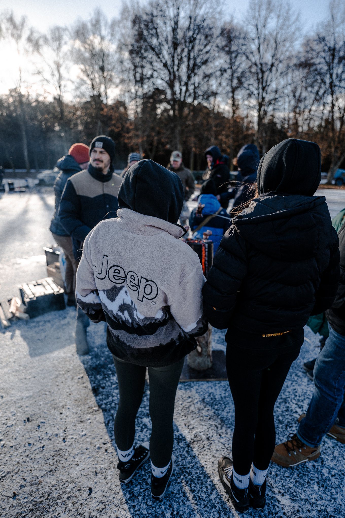 Group of people dressed in winter clothing gathered outdoors on a snowy ground, with leafless trees in the background.