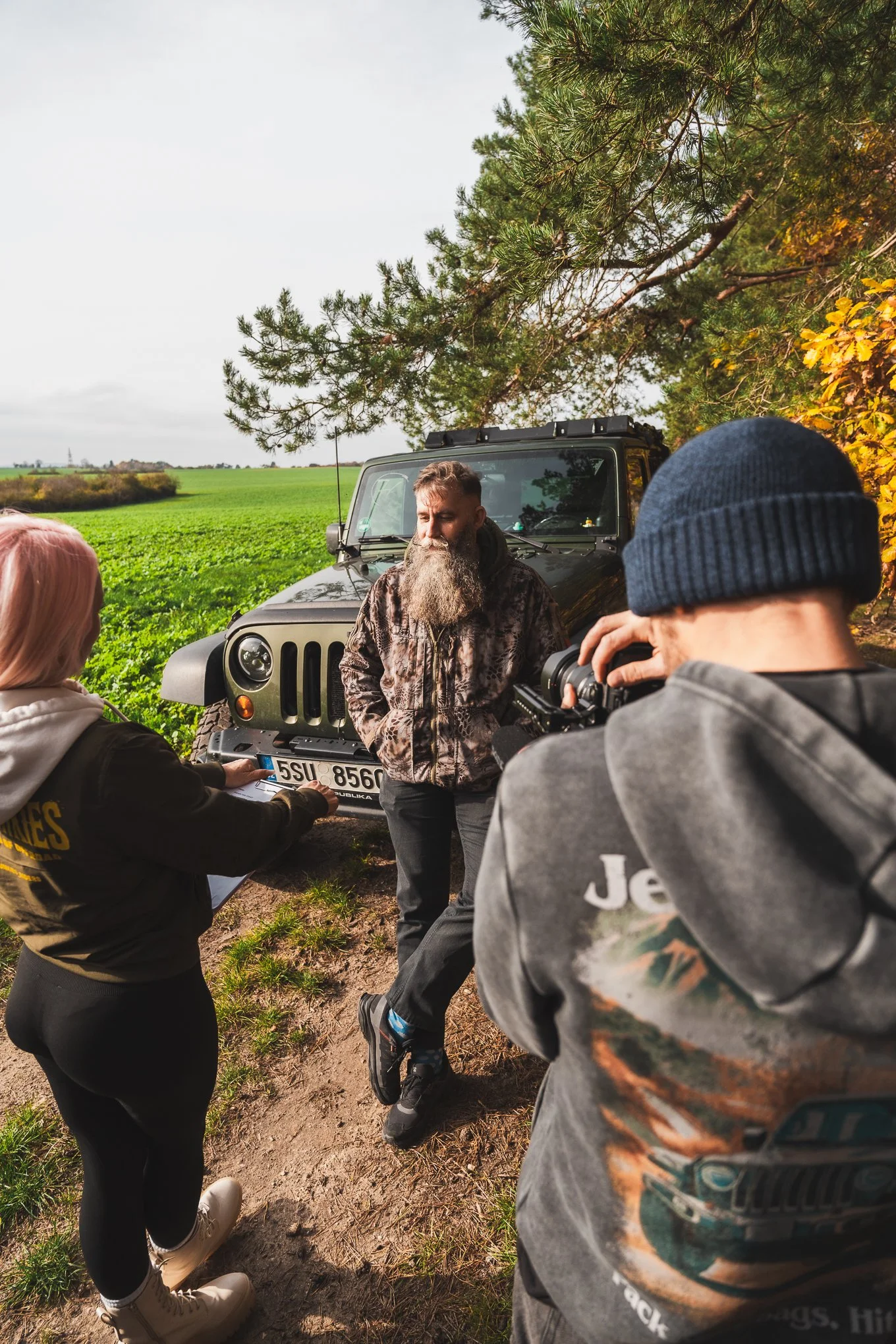 A man with a long beard in camouflage jacket is being filmed or interviewed by a woman while a photographer captures the scene. They are outdoors near a Jeep, with green fields and trees in the background.