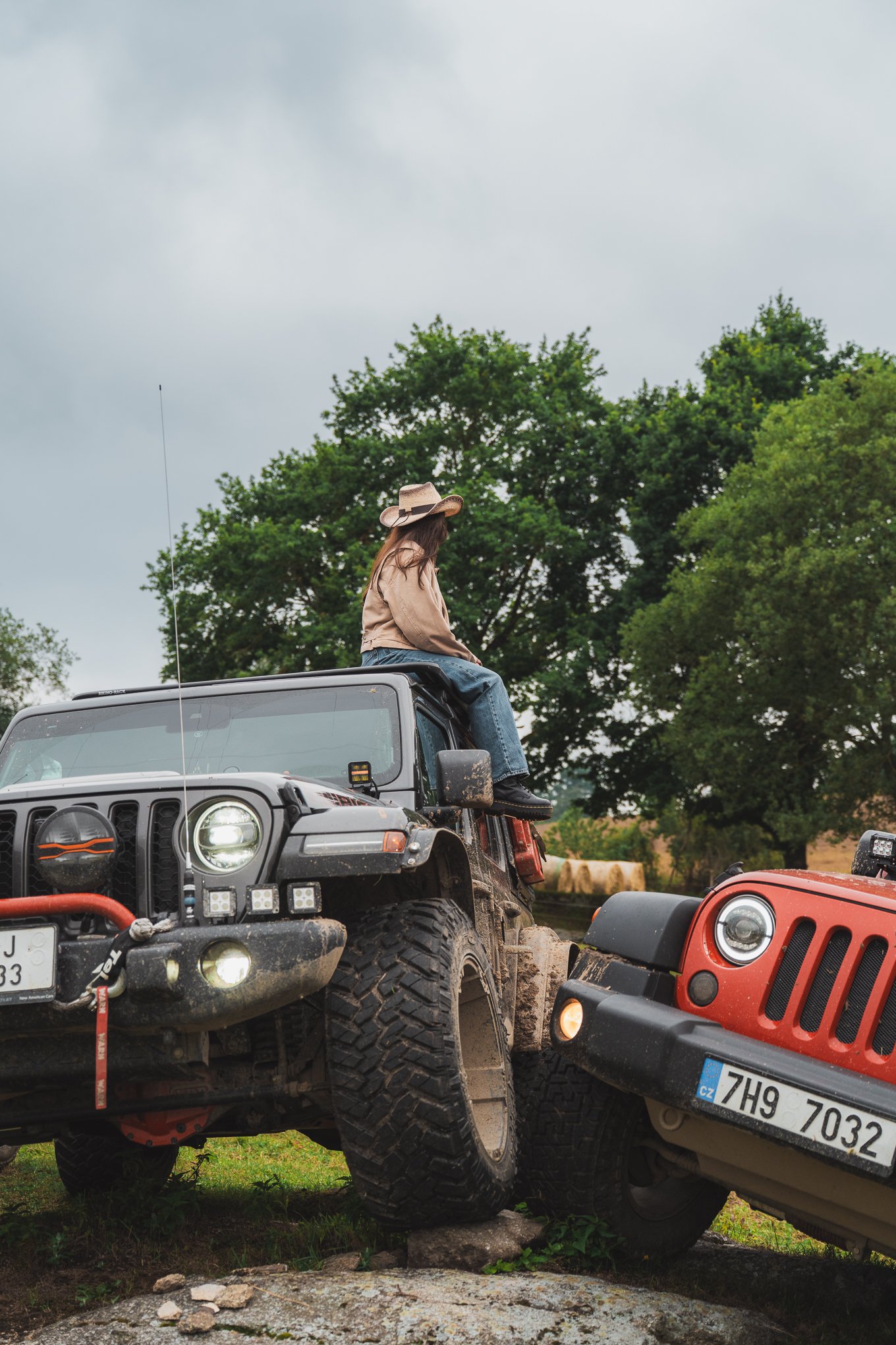 A woman sitting on the roof of a black Jeep with a woman sitting on it, next to a red Jeep, on a rocky terrain with trees in the background.