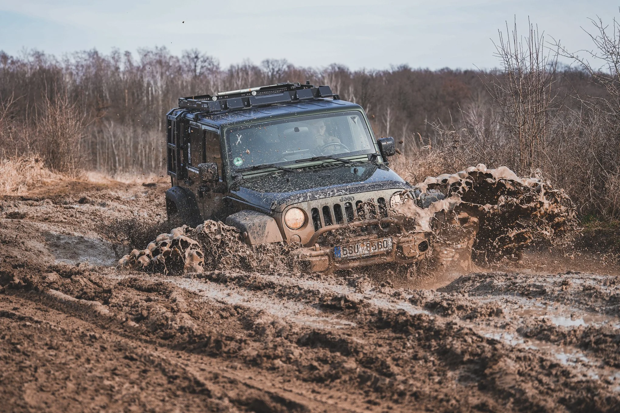 A black Jeep vehicle driving through muddy off-road terrain, splashing mud and water.