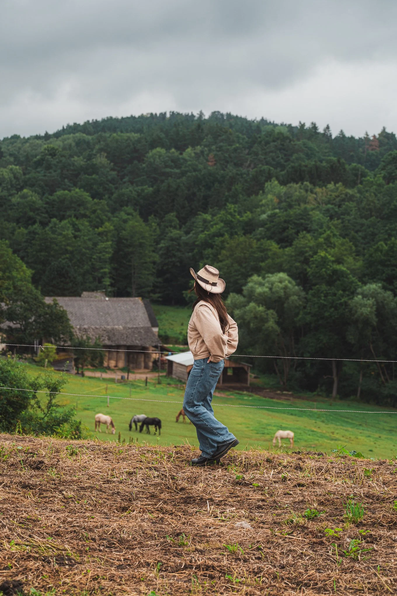 A woman standing on a rural hillside with her hands in her pockets, wearing a beige jacket and wide-brimmed hat, looking at a farm with horses grazing, surrounded by green trees and mountains under cloudy skies.
