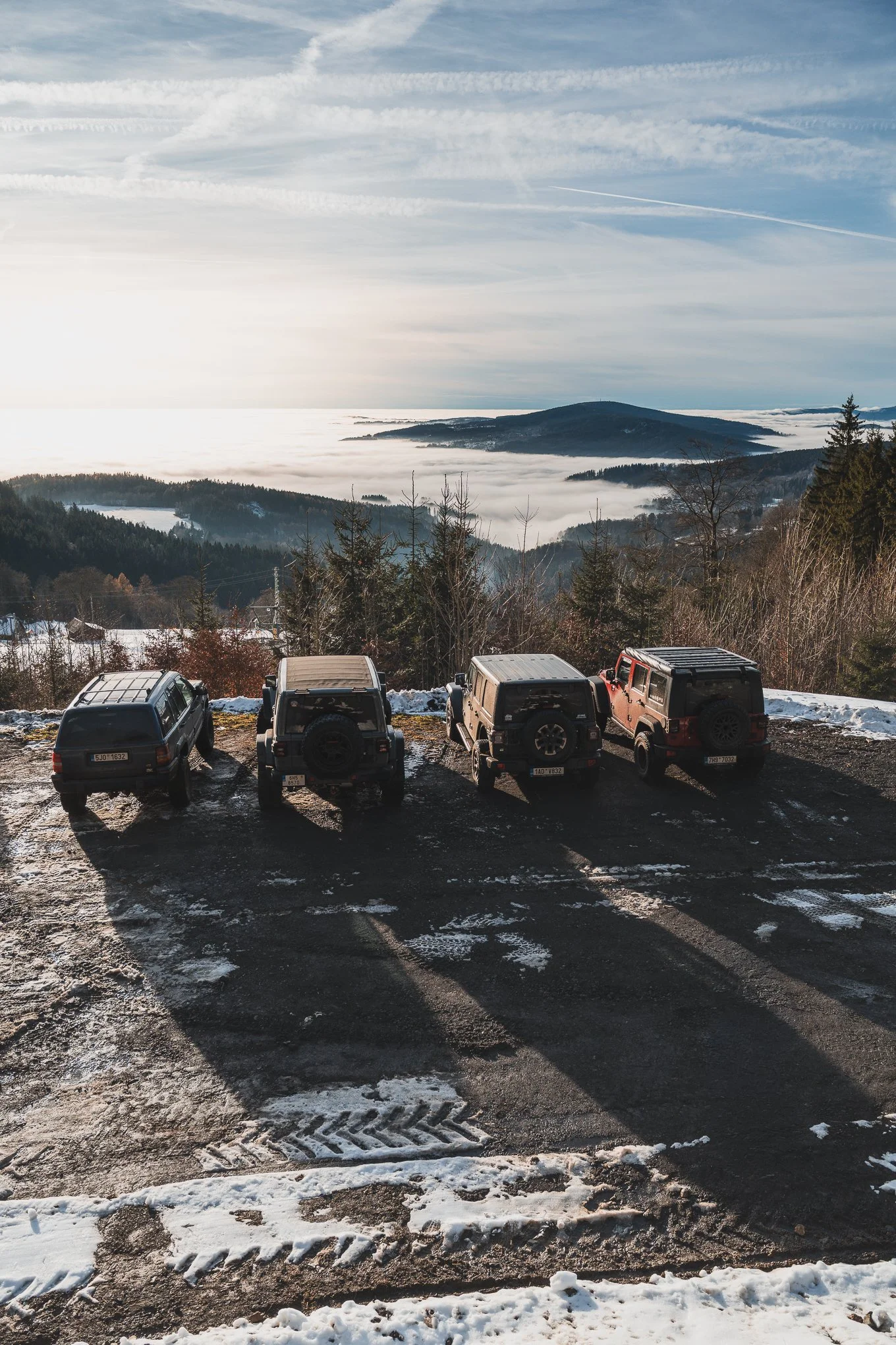 Four parked vehicles in a snowy mountain overlook with a scenic view of clouds and mountains in the background.