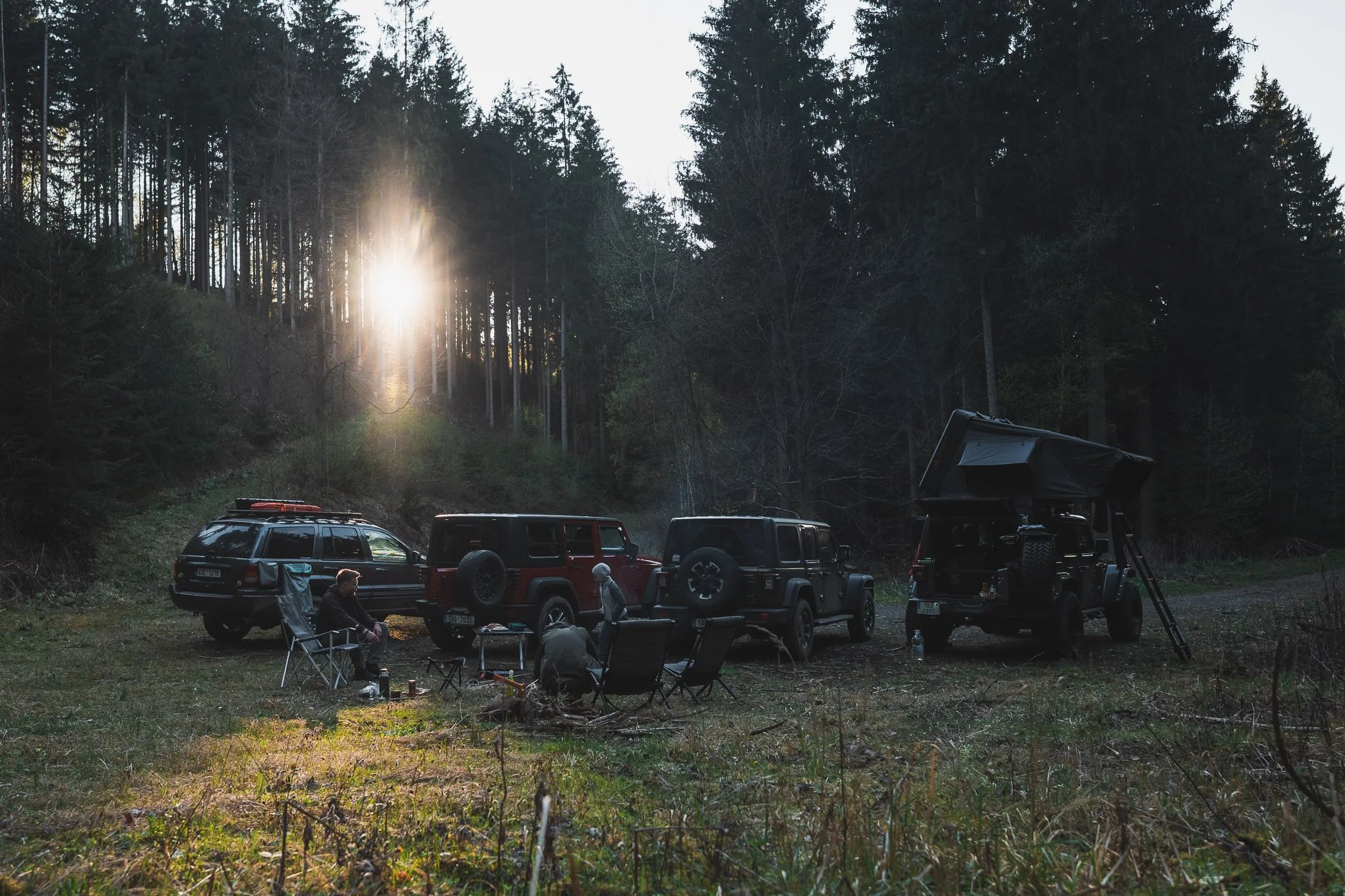 Four vehicles parked in a clearing near a forest, with a group of people sitting and standing around camping chairs and a table, as the sun sets through tall trees.