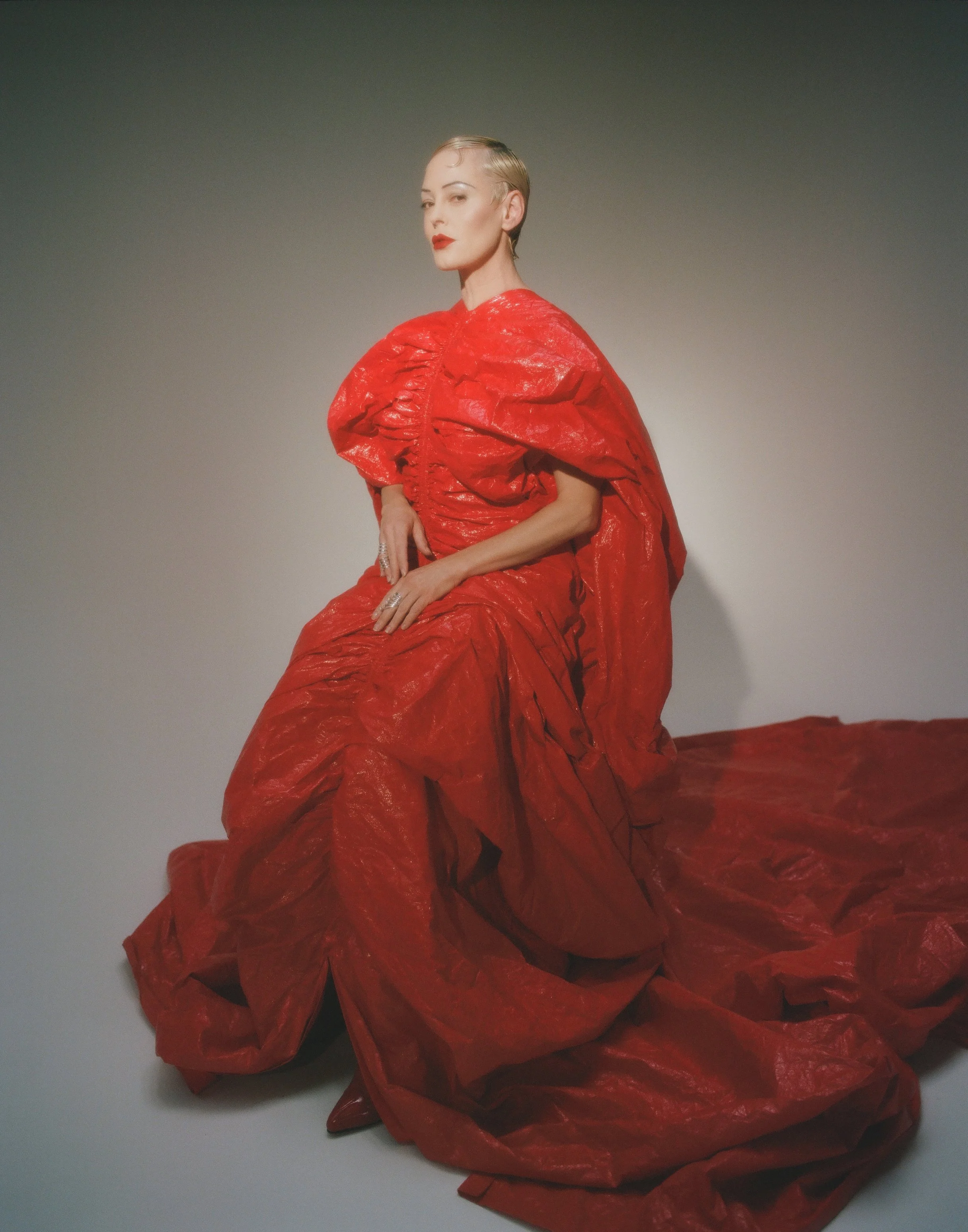 Rose Mcgowan in a striking red, textured, and dramatic gown poses against a plain background. Shot by Timo Kerber